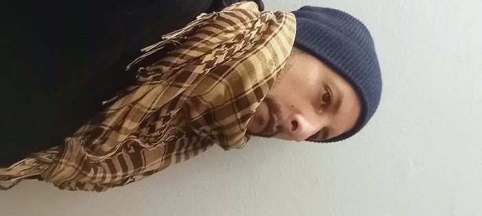 A man wearing a blue knit hat and a brown and beige checkered scarf stands against a white wall. He is wearing a black shirt or jacket. The man looks down and slightly to the right. The scarf is draped over his neck and partially covers his face. The image is rotated slightly counter-clockwise.