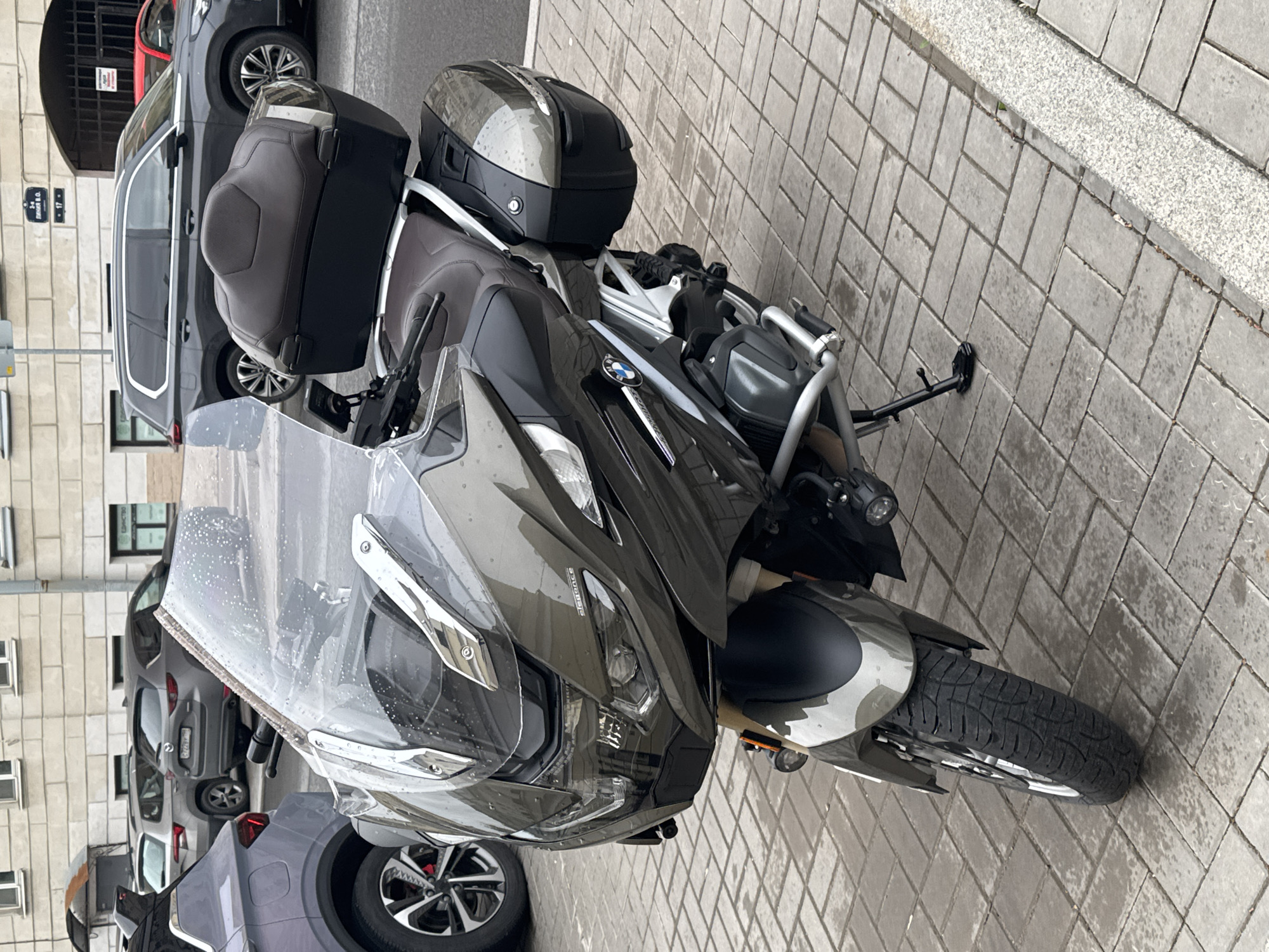 The image depicts a BMW R 1250 RT motorcycle parked on a sidewalk. The motorcycle is equipped with top cases and a front windshield. The location is Saint Petersburg, Russia, as indicated by the street sign "Яковлевский пер. 17" (Yakovlevsky pereulok 17) visible in the background. Several cars are parked along the street and an apartment building is in the background. The motorcycle is parked on a paved sidewalk.