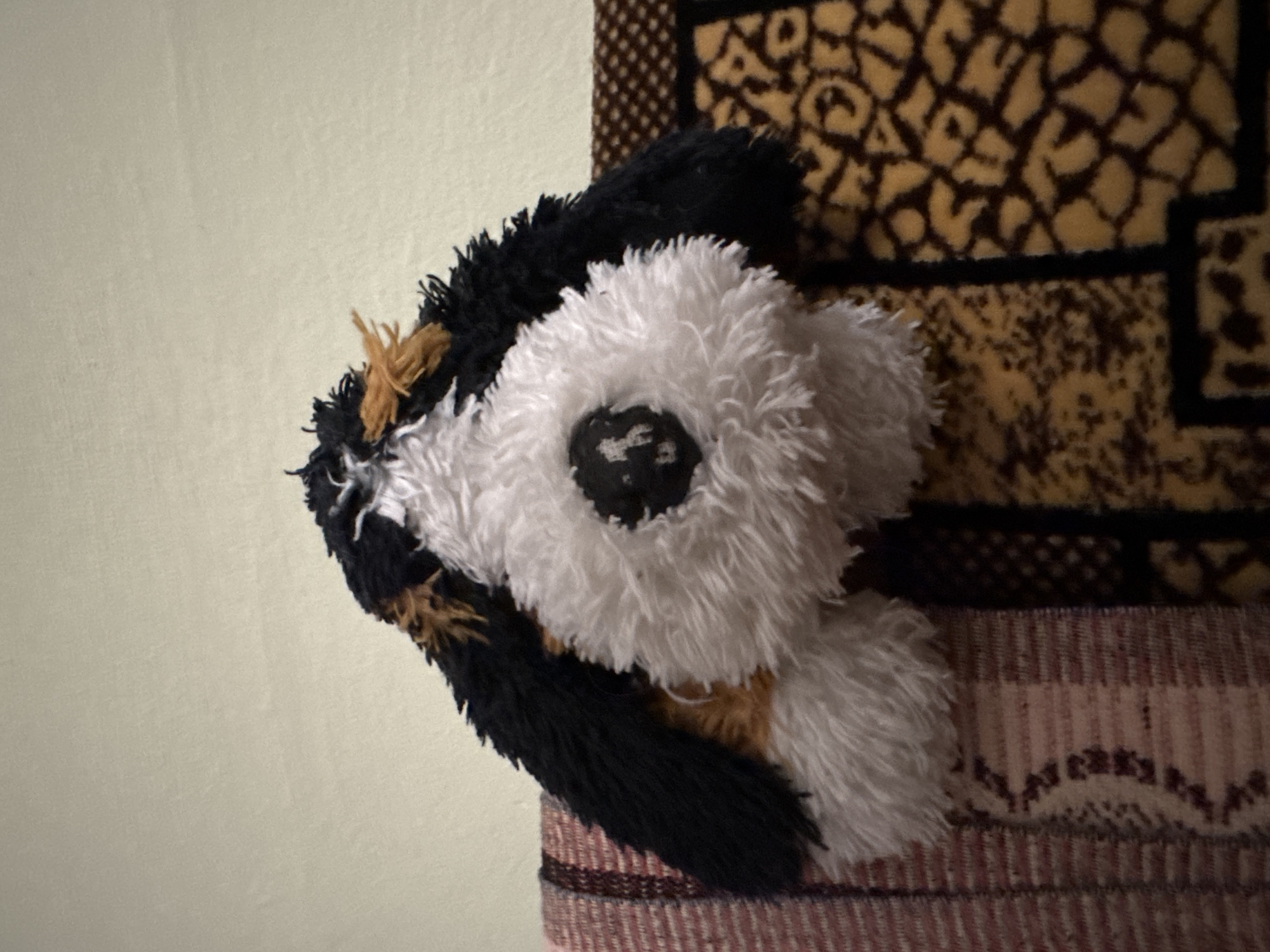 An overhead, medium shot captures a fluffy, plush toy dog in dark brown, black, and white fur. The toy is positioned centrally in the frame, resting on a patterned fabric surface. The dog's head is tilted downward, obscuring its face, but a black, circular nose is visible, with some white stitching details on its surface. The dog's body is predominantly white and fluffy, with patches of black and brown fur around its head and ears, giving it a tri-colored appearance. It appears to be sitting on a sofa or armchair, with the patterned fabric beneath it displaying a combination of warm tones, including yellow, brown, and black, in an abstract or animalistic print. To the left of the toy dog, a different fabric is visible, featuring a striped pattern in shades of pink, white, and dark red. The background is a plain, light-colored wall, creating a simple backdrop that emphasizes the toy and its immediate surroundings. The lighting is dim, casting soft shadows and highlighting the textured surfaces of the plush toy and the fabrics. There are no people or discernible actions in the image.
