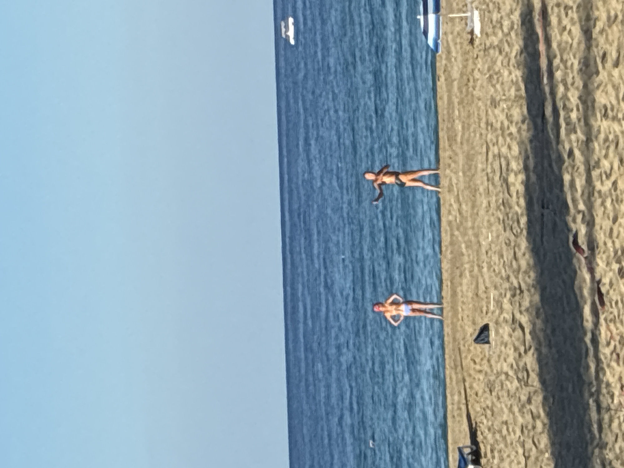 In Alanya, Turkey, on a sandy beach, two individuals are visible near the waterline under a clear blue sky. The person on the left, wearing a pink headscarf and a light blue bikini, stands with hands on hips, facing forward. To the right, another person in dark swimwear faces left, with their right arm extended in a gesture as if throwing something. The sea is a deep blue, with a white boat visible in the distance. The foreground shows the textured sand of the beach, with shadows cast by unseen objects, and a blue and white striped umbrella is partially visible on the right side of the frame.