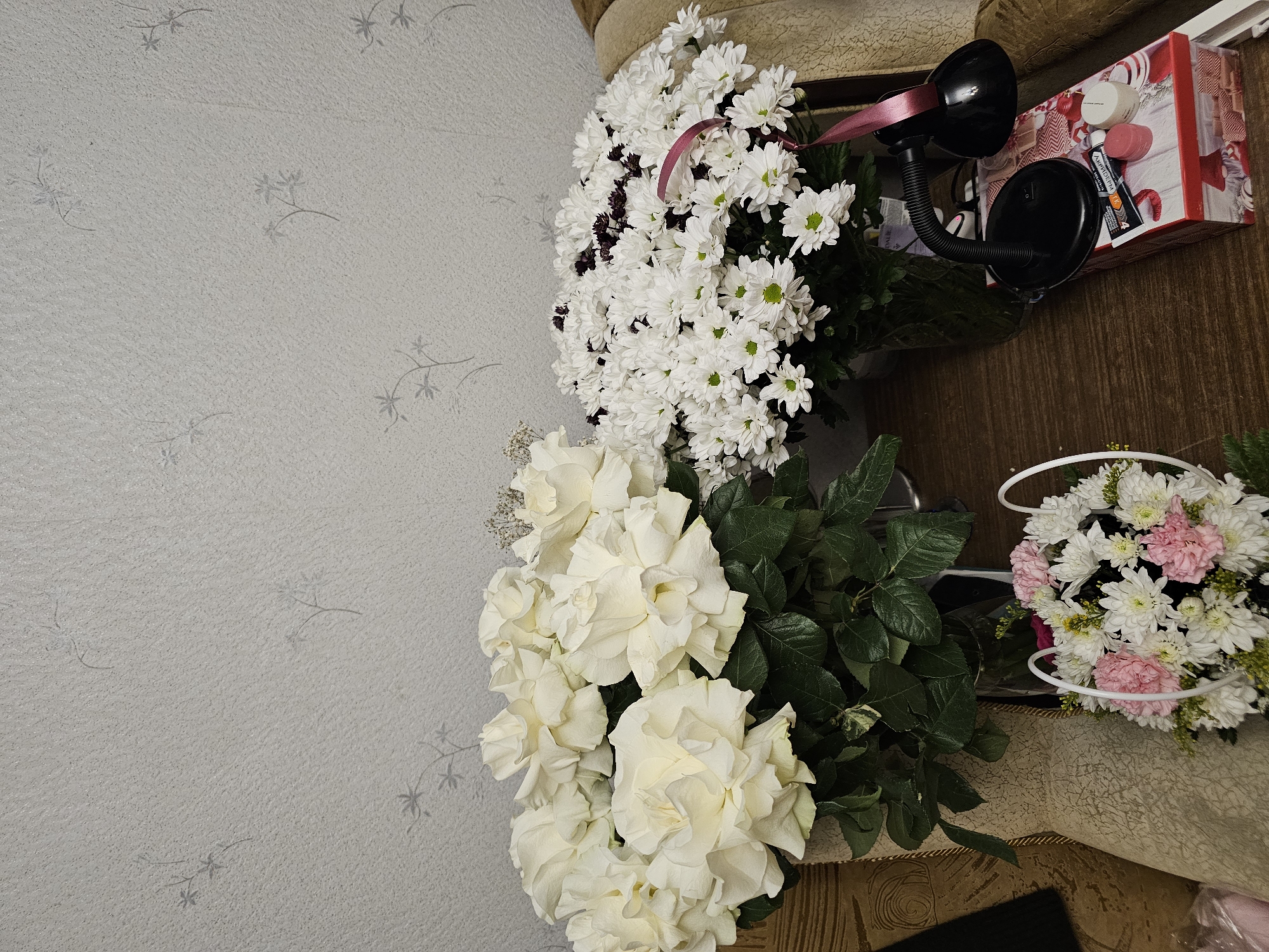 The image shows a scene in Khimki, Russia, featuring three bouquets of flowers. One bouquet consists primarily of white roses with accompanying green foliage. Another bouquet is a mix of white and darker purple flowers. The third bouquet contains white and pink flowers. A small desk lamp and a box with various beauty and personal care products are also visible. The backdrop includes a patterned wall and a portion of what appears to be a couch.