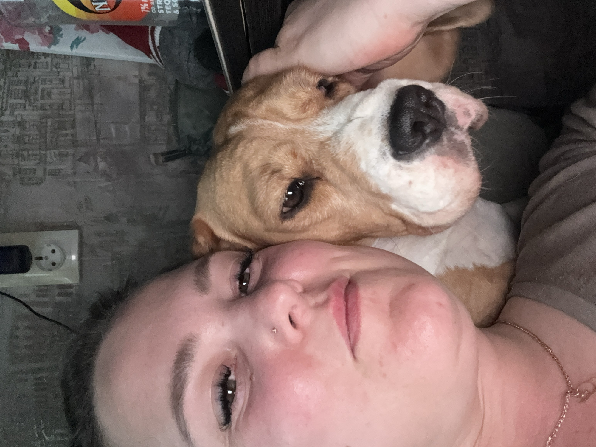 A woman is lying down with a dog. The dog is a tan and white color and has its head resting on the woman. The woman has dark hair and a nose ring. A power outlet is visible on the wall behind them. The city location, Марковский, Russia, is not visually discernible from the image.