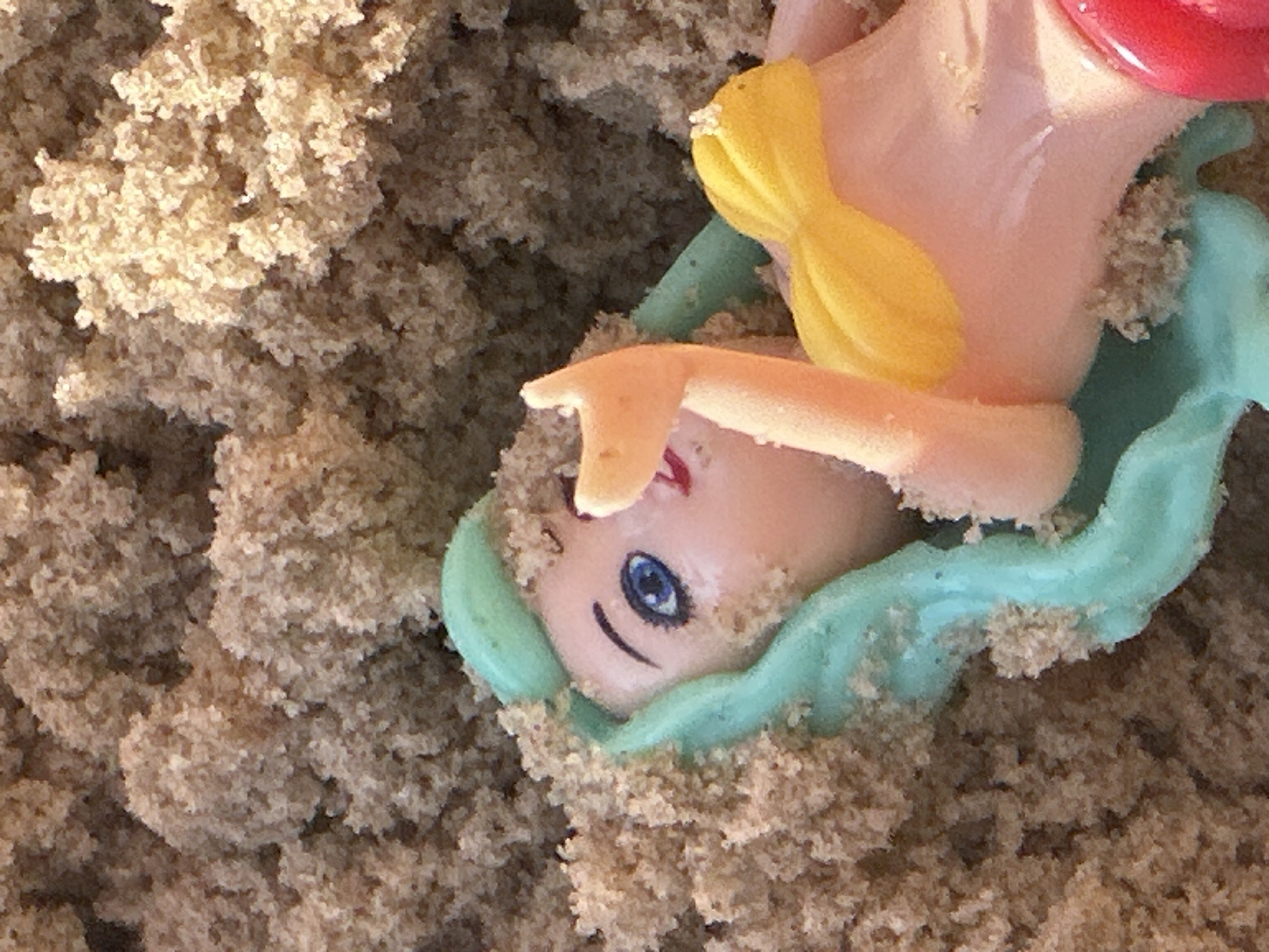 A doll with blue eyes and turquoise hair is partially buried in sand. The doll is wearing a yellow bikini top and a red bottom. The scene appears to be a beach in Herzliya, Israel.