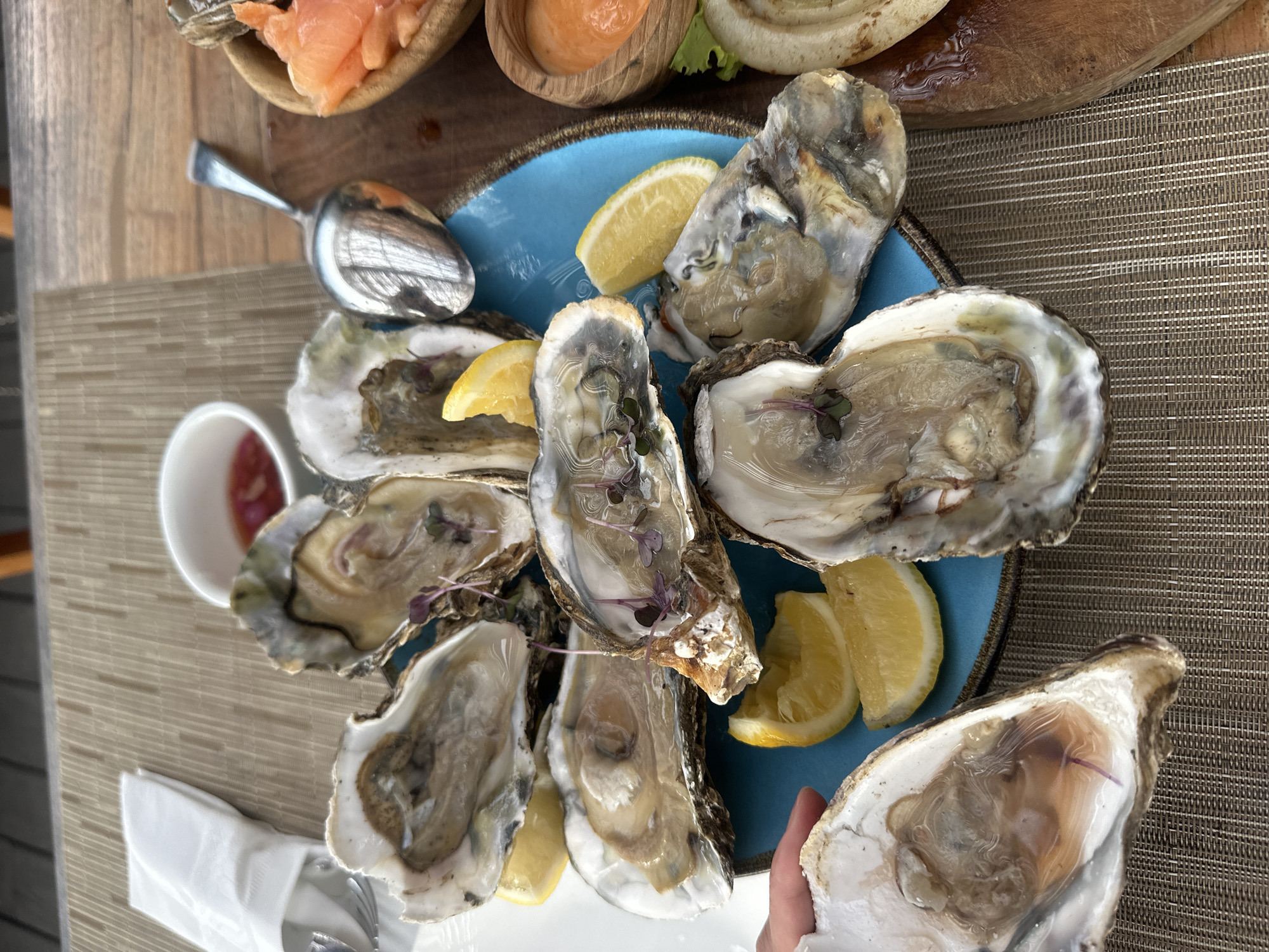 The image shows a plate of oysters on a table in North Kuta, Indonesia. The oysters are served with lemon wedges and small sprigs of herbs. A small bowl of sauce is visible nearby. Other food items, possibly salmon and condiments, are in the upper left corner. A spoon is next to the plate. The table has a textured woven surface. A person's hand is partially visible in the lower right corner.