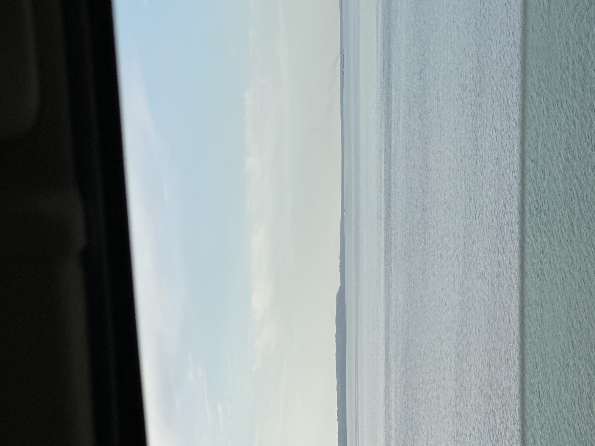 The image shows a body of water, likely the Kama Reservoir in Chaikovsky, Russia, with land visible in the middle ground. The sky is overcast, and the image is taken from inside a vehicle, as indicated by the frame on the left side of the photo. The water's surface is textured with waves or ripples, indicating wind or current.