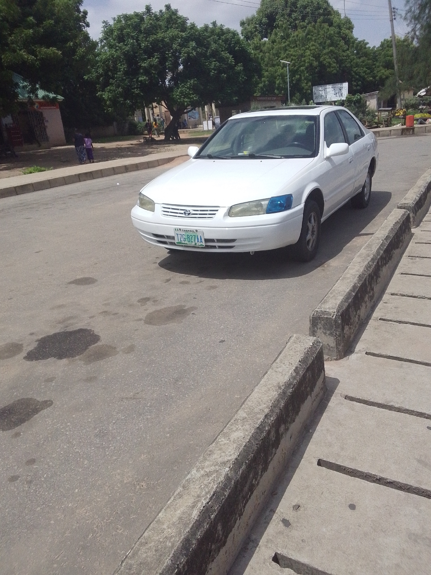A white Toyota Camry sedan is parked on an asphalt road in Jalingo, Nigeria. The car's license plate reads "TZG-827AA" and displays "TARABA" at the top. The car's headlights have a distinctive blue tint. In the background, several large trees with lush green foliage line the street, and a few buildings are visible. A concrete curb with drainage channels runs along the right side of the road. The scene appears to be set during the daytime, with bright sunlight casting shadows on the ground, suggesting fair weather. Two people, one in a dark outfit and the other in a light purple top and patterned skirt, are walking on the sidewalk in the middle ground, near a building with a green roof. Further in the distance, more people are visible near other structures, suggesting a community setting. The overall impression is a quiet street scene in a Nigerian town.