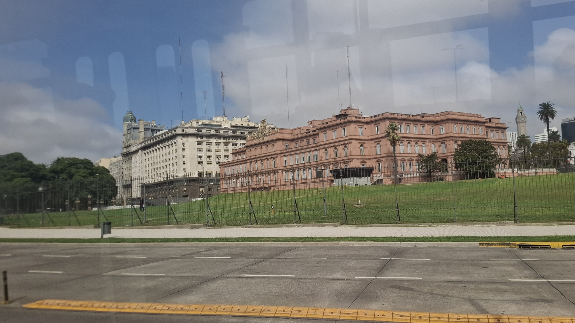 The image shows the Casa Rosada, the executive mansion and office of the President of Argentina, located in Buenos Aires. The building is pink and is situated behind a green lawn and a black fence. To the left of the Casa Rosada is a white building with multiple stories and a green-domed tower. In the background, a clock tower and palm trees are visible. In the foreground, there is a gray road with white lines and a yellow speed bump. The image appears to be taken from inside a vehicle, as evidenced by the reflections in the glass. The sky is blue with scattered clouds.