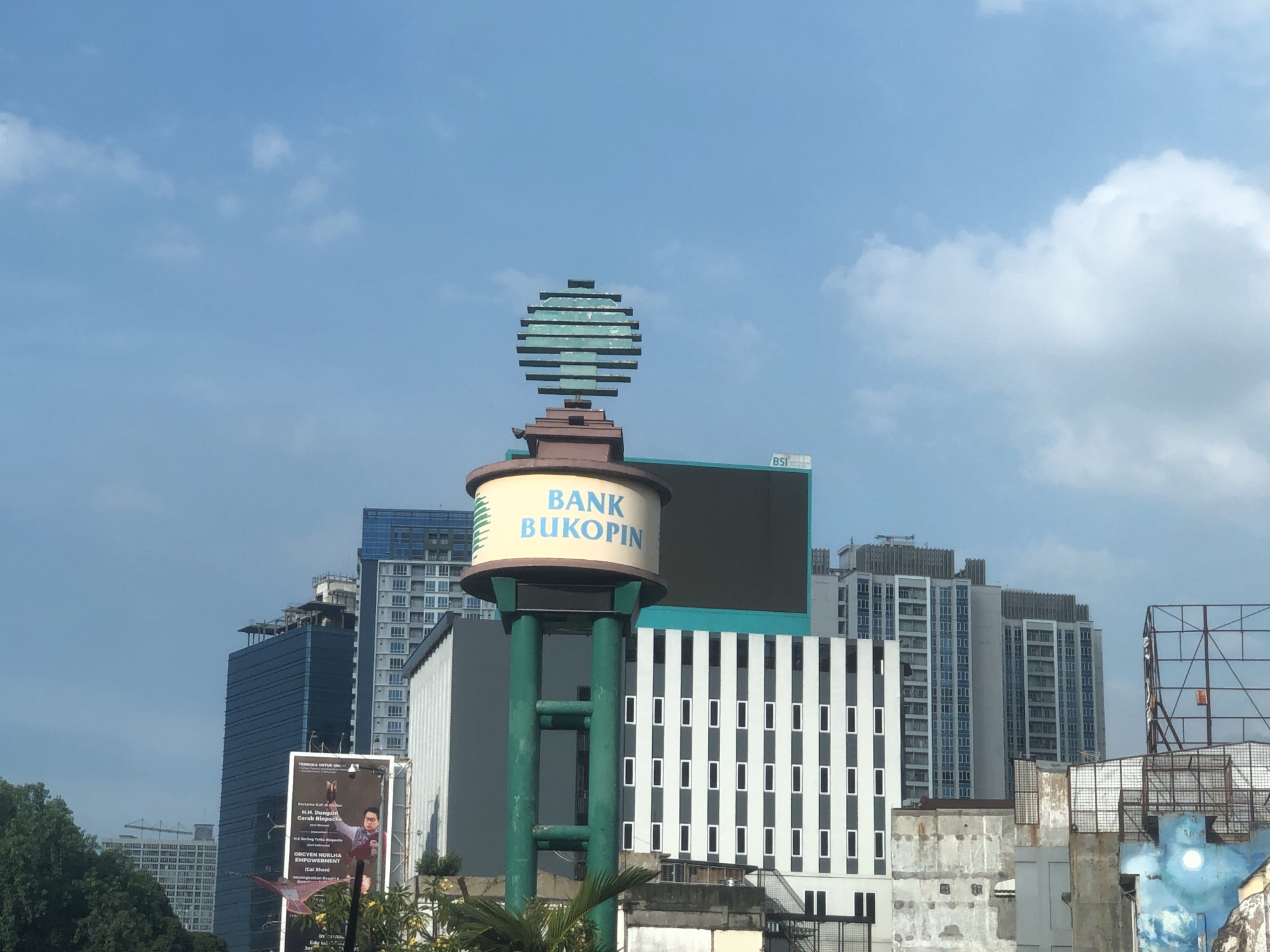 The image captures an urban landscape in the City of Medan, Indonesia. A prominent feature is the Bank Bukopin sign situated on a high structure, with a stylized spherical decoration on top. Surrounding the sign are various buildings, including modern high-rise structures with multiple windows and contrasting white and dark facades. A billboard featuring a portrait and text is visible on the left side of the frame. The sky is blue with scattered clouds. The overall atmosphere suggests a daytime urban setting with a mix of commercial and residential buildings.