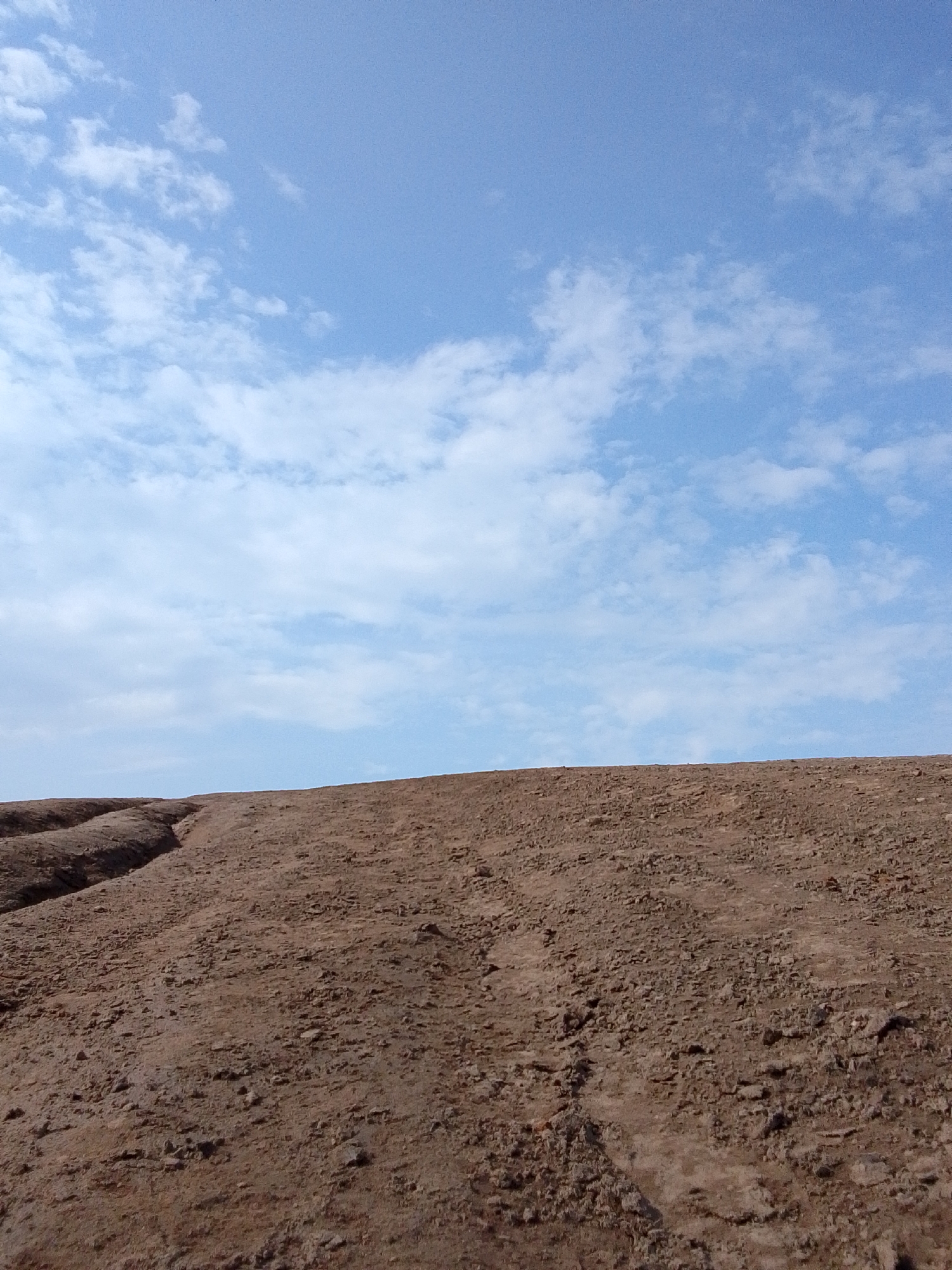 The image shows a landscape under a partly cloudy, blue sky. The lower portion of the image is dominated by reddish-brown soil, possibly dirt or a mound of earth. The soil appears dry and consists of small stones or pebbles. There is an eroded channel or ditch cutting through the soil on the left side.
There are no visible people, animals, or buildings. The location is assumed to be in or near 1-Go Maya, Russia, based on the prompt's information. The scene lacks other specifics, like vegetation, infrastructure, or structures.