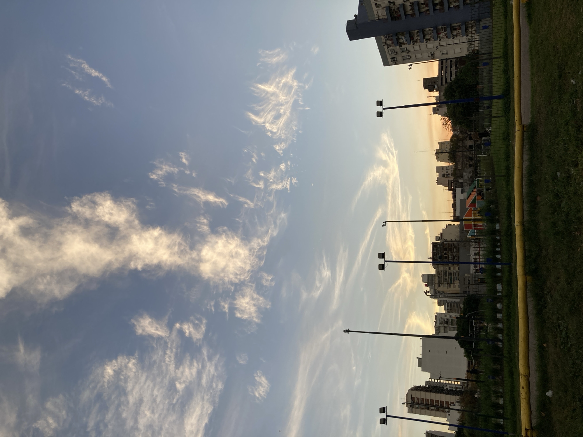 The image depicts a sky with wispy clouds above a cityscape, possibly captured during sunrise or sunset. The location is speculated to be in Buenos Aires, Argentina, based on the architectural style of the buildings. The scene includes tall apartment buildings, streetlights, a grassy area along the edge, and a glimpse of playground equipment. No people are visible in the image. The lighting suggests a time of day with warm hues, potentially dawn or dusk.
