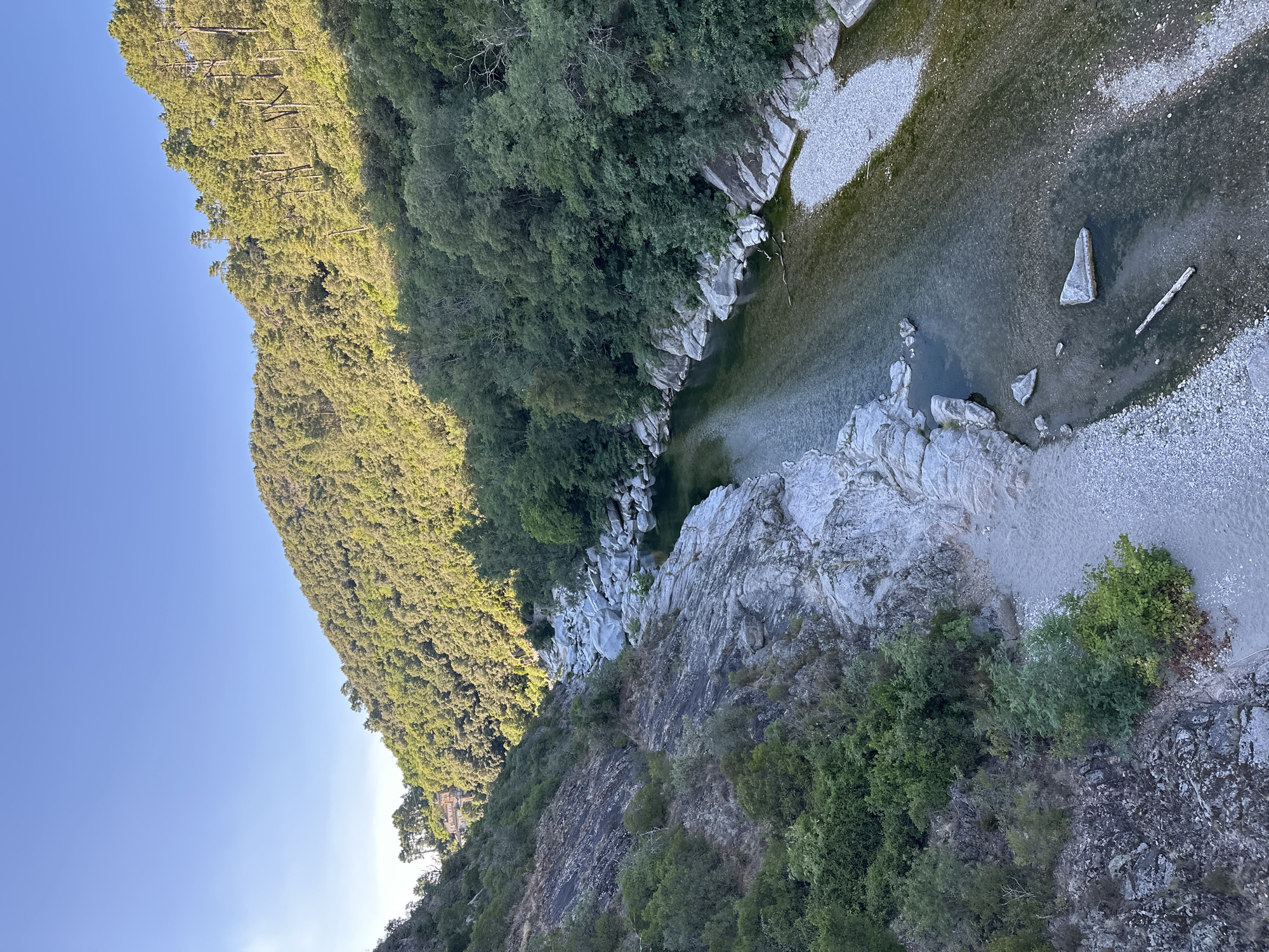 The image depicts a serene natural landscape featuring a river flowing through a rocky gorge. The riverbanks are lined with a mix of lush green trees and grey, weathered rocks. Sunlight illuminates the scene, casting shadows and highlighting the textures of the natural elements. In the distance, a red-roofed building is partially visible nestled on a hillside covered with dense foliage. The sky above is a clear, bright blue. This vista is likely located in the vicinity of Anduze, France, a town known for its natural beauty and proximity to the Cévennes National Park, which features similar rugged terrain and winding rivers.