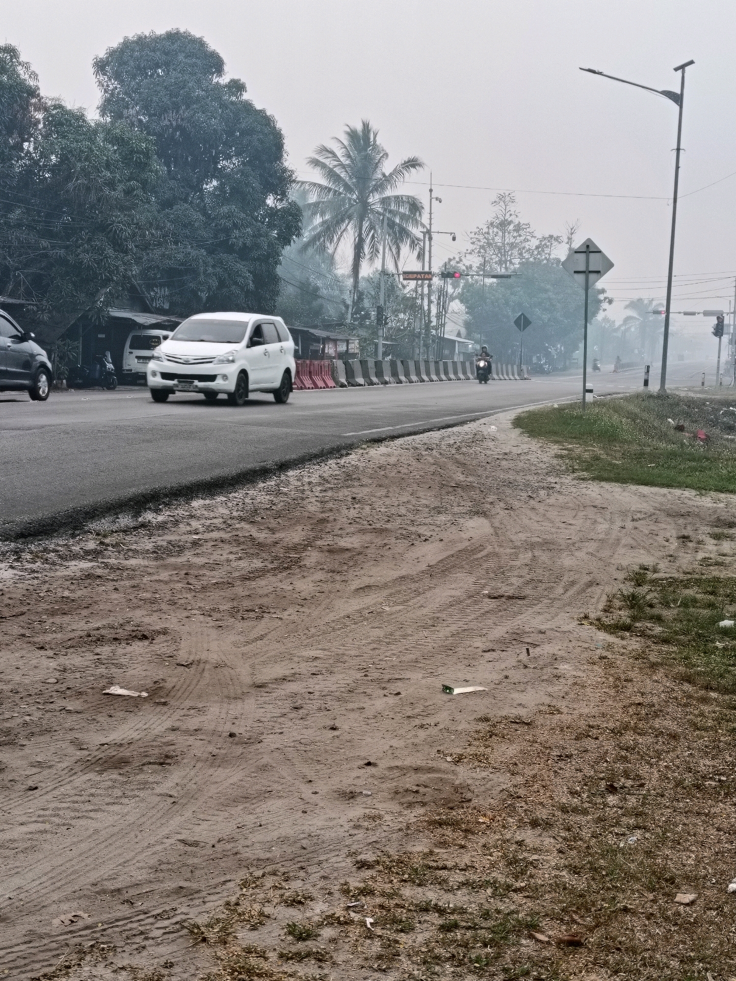 A wide, paved road runs through a hazy, overcast environment, likely during the day in Palangka Raya, Indonesia, given the presence of a prominent palm tree and the general atmosphere. The road appears to be undergoing some construction or repair, with loose dirt and tire tracks on the shoulder in the foreground. Several vehicles are present: a white minivan in the middle lane, a dark-colored car in the left lane, and a motorcycle rider further down the road. Traffic lights are visible, with one displaying a red light. A lamppost and various road signs are also present. Vegetation, including trees and grass, borders the road. The overall impression is one of a slightly overcast, industrial or developing area on the outskirts of a city.