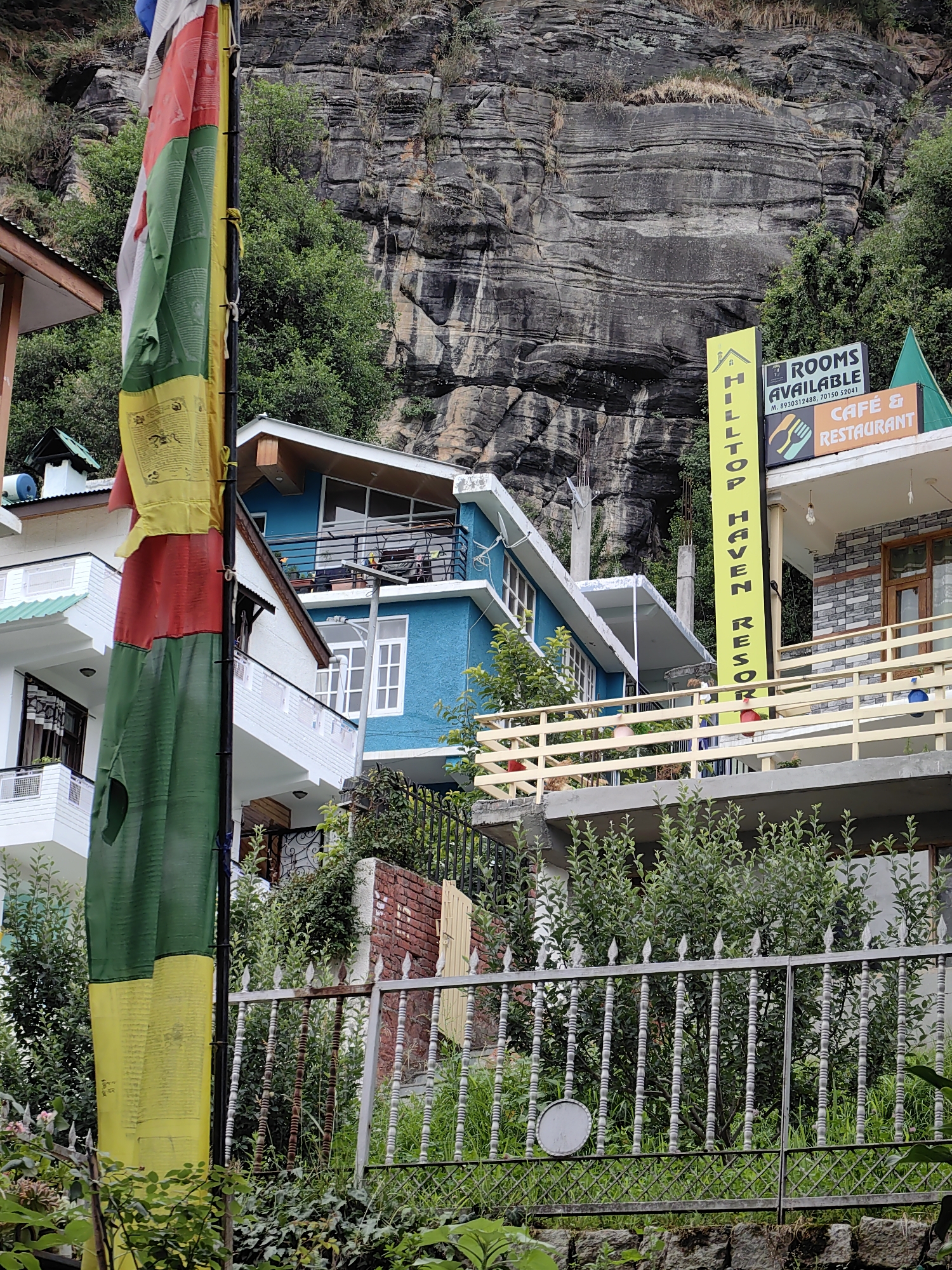 The image shows a street scene in Vashisht, India, with buildings nestled against a rocky mountainside. A prayer flag, featuring the colors red, green, yellow, and white, hangs prominently in the foreground, partially obscuring the view of the buildings. To the right of the flag, a yellow vertical sign reads "HILLTOP HAVEN RESORT" and a smaller sign indicates "ROOMS AVAILABLE." Below this, an orange sign reads "CAFE & RESTAURANT." The buildings visible include a blue structure with balconies and a white structure with a green roof. A metal fence with decorative finials runs across the lower portion of the image, separating the street from a garden area. Lush green foliage and trees are scattered throughout the scene, with the rugged, layered rock face of the mountain dominating the background. The sky is overcast, suggesting a cloudy or rainy day. No people are visible in the image.