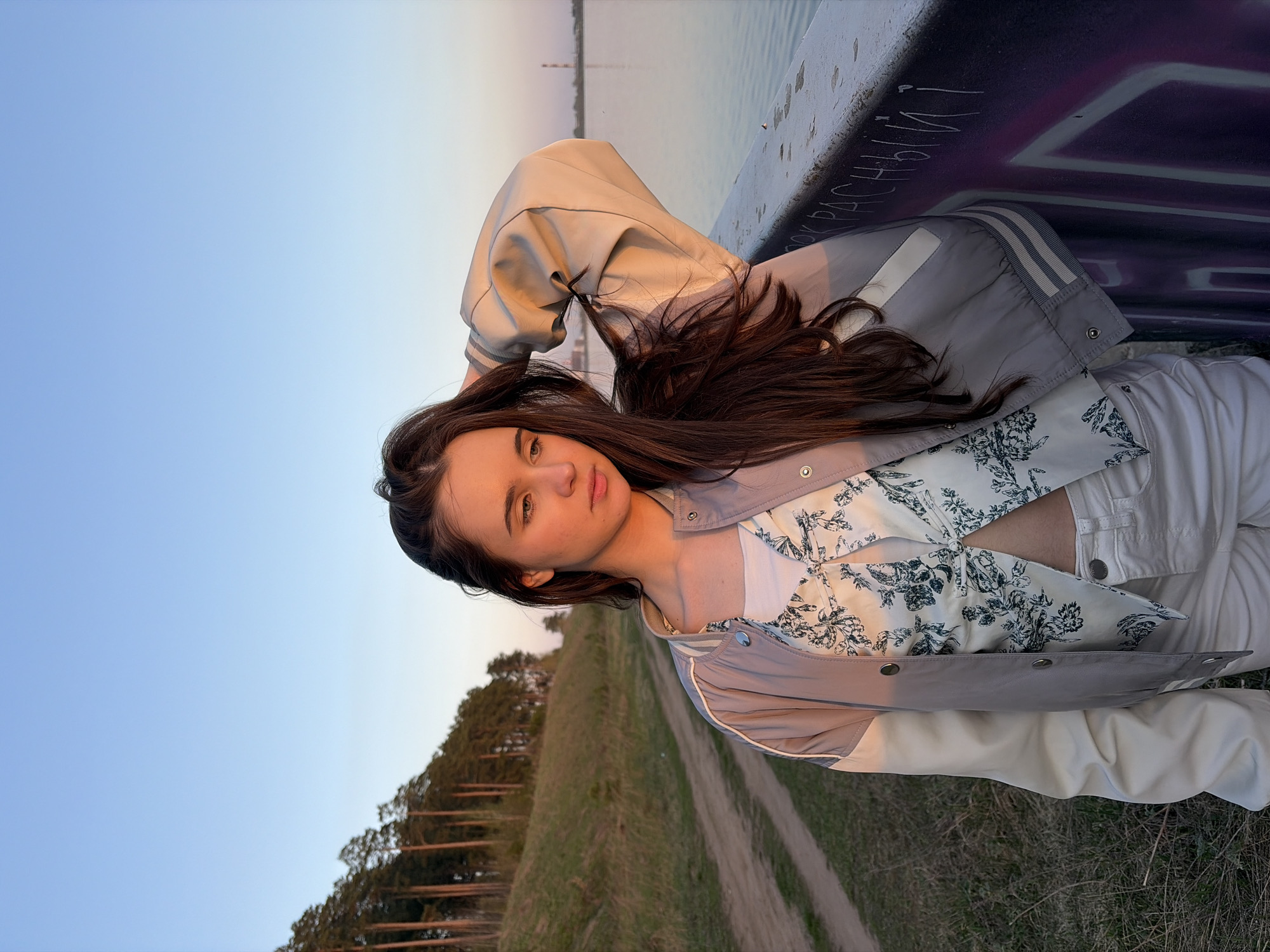 A young woman is lying on a ledge with graffiti, possibly a bridge in Izhevsk, Russia, based on the landscape. She is wearing white pants, a floral print top, and a beige and grey jacket. The background shows a river, trees, a path, and a gradient sky transitioning from blue to pink.