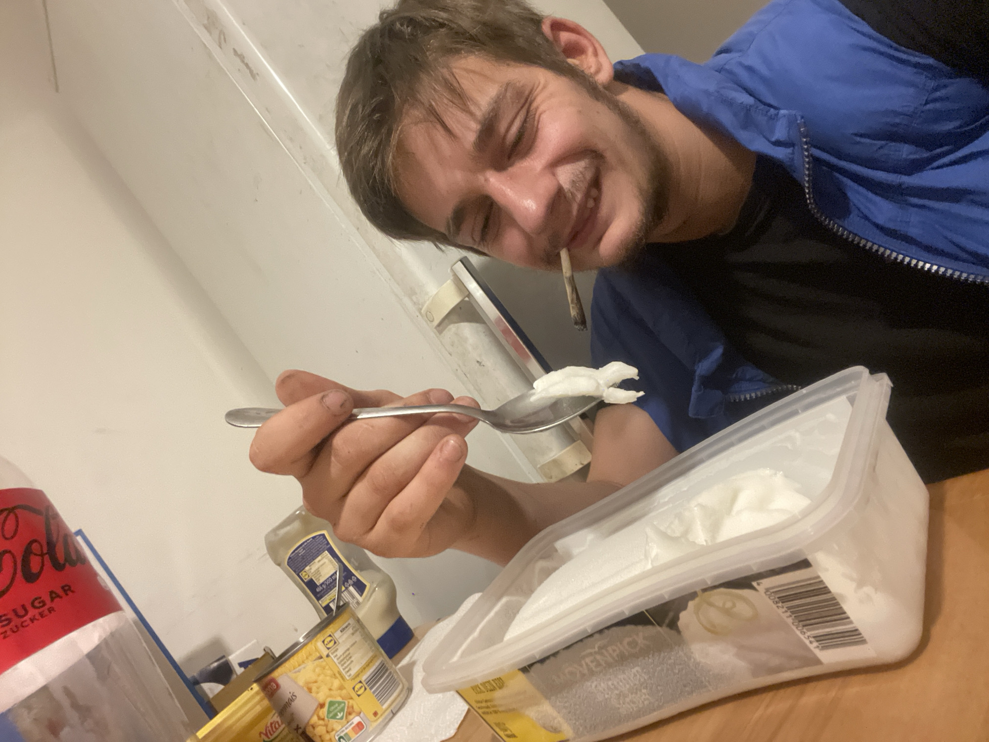 The photograph shows a young man with short brown hair and a stubbly beard, wearing a blue jacket over a black shirt. He has a lit joint in his mouth, and he's smiling at the camera. He holds a spoon with a dollop of white cream in his right hand. Beside him on a wooden surface are a transparent plastic container filled with more white cream, a bottle of mayonnaise, a can of corn, and a large bottle of Cola Sugar. The background consists of a plain white wall. The location appears to be inside a residential home, but the city is unknown.