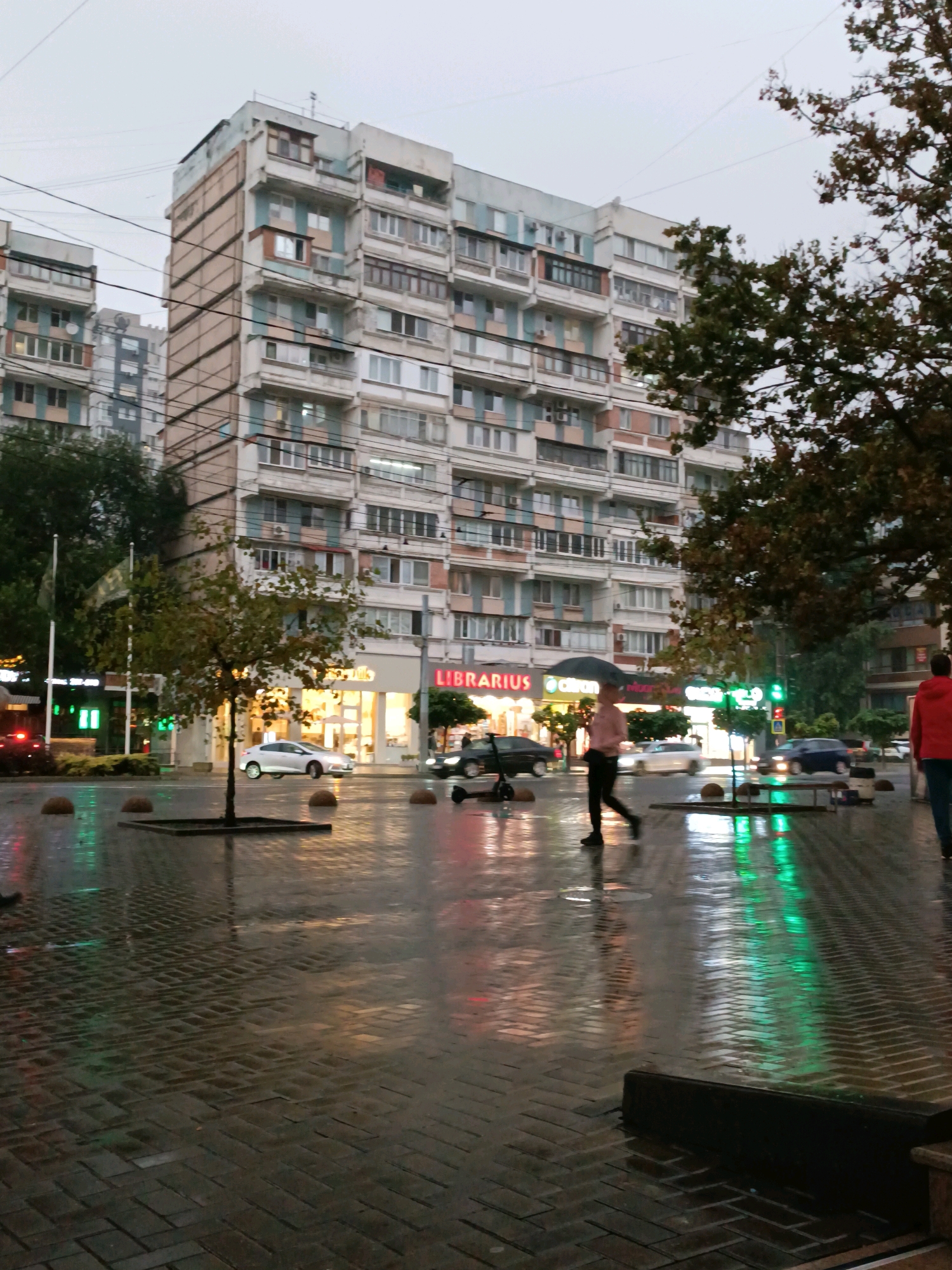 The image depicts a street scene in Chișinău, Moldova, on a rainy day. A large, multi-story apartment building with a beige and light blue facade dominates the background. The building features numerous windows and balconies, with some windows appearing to be illuminated. In front of the building, a row of shops lines the ground floor, one of which displays the sign "LIBRARIES".
The foreground shows a wet, paved sidewalk reflecting the ambient light. A person with an umbrella is walking across the street, and another person in a red jacket is visible on the right side of the frame, also walking. Several cars are present on the wet street, some of which are moving, creating reflections on the surface. A small tree with sparse leaves is planted in a square planter in the middle of the sidewalk. The overall atmosphere is subdued, indicative of overcast and wet weather conditions.