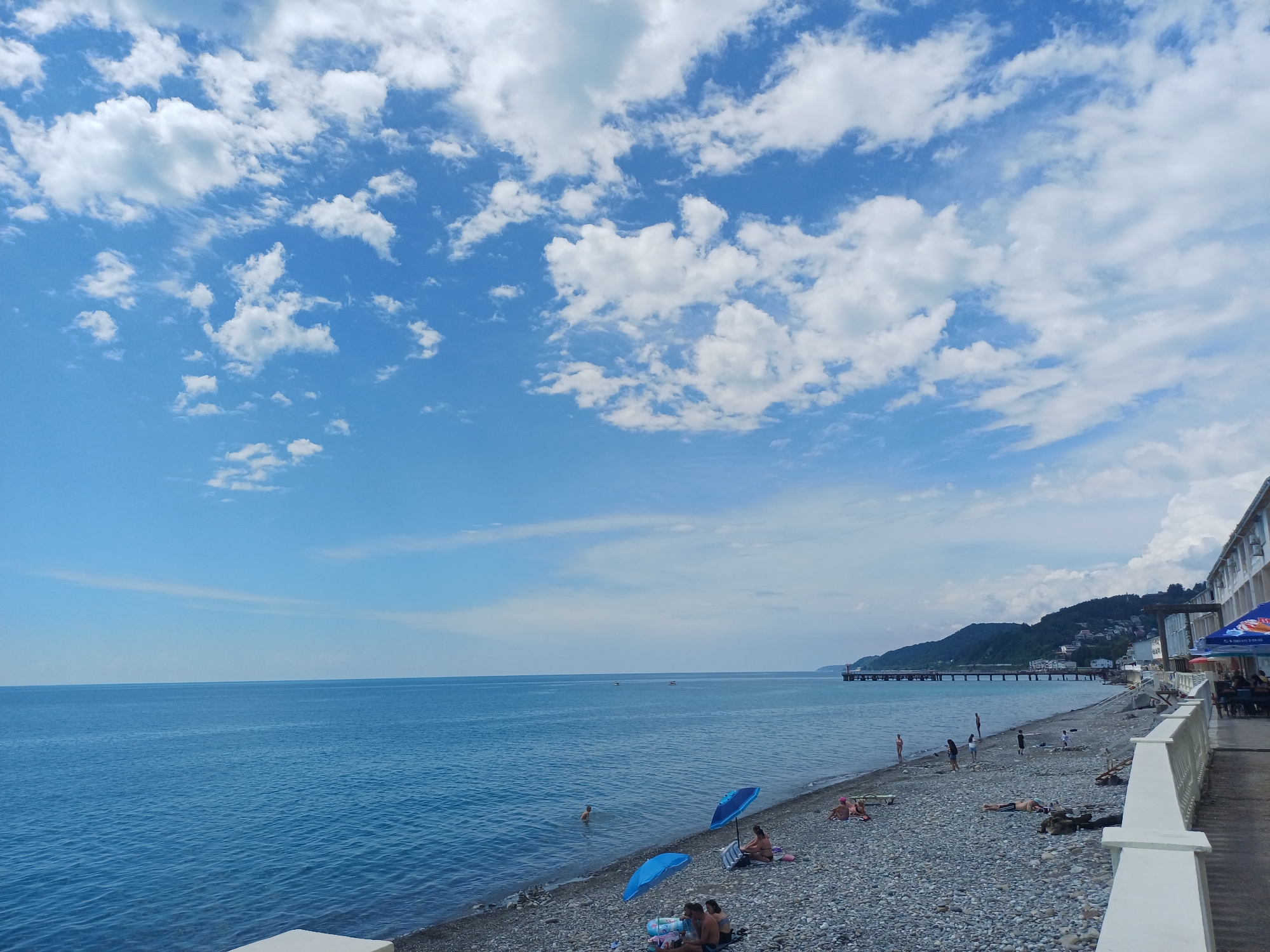 The image depicts a coastal scene with a pebble beach bordering a calm, blue sea under a partly cloudy sky. On the beach, several people are visible, some sunbathing, others sitting under umbrellas, and a few appear to be swimming in the water. There is a building with a white railing to the right of the frame, and a long pier extends into the sea in the distance, backed by a tree-covered hillside with scattered buildings. The overall impression is of a resort area, likely Sochi, Russia, given the architectural style and mountainous backdrop.