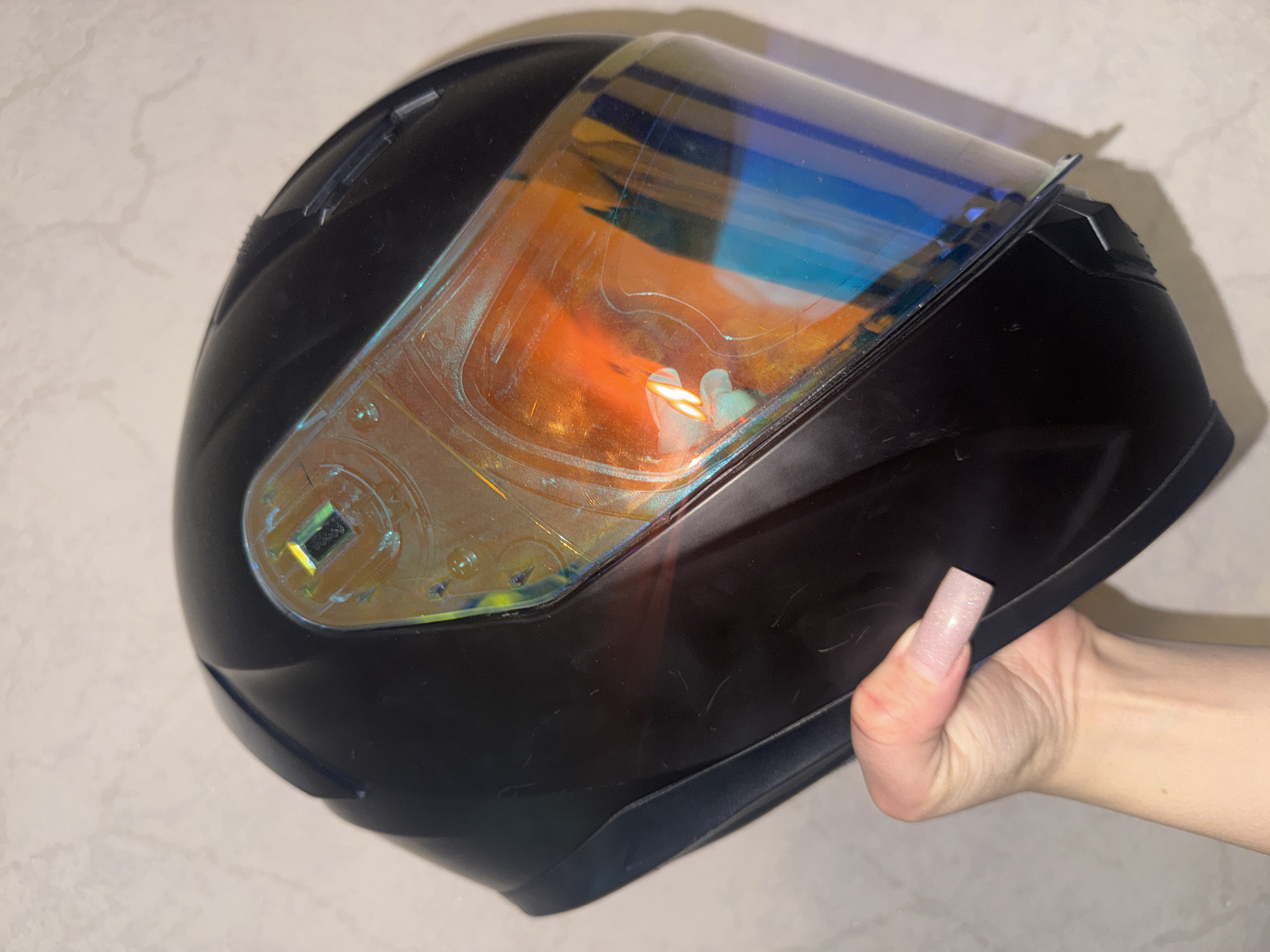 A black motorcycle helmet with a reflective, iridescent visor is held in a person's left hand. The hand has long, square-shaped, light pink acrylic nails. The helmet is positioned at an angle, with the visor facing the camera. The background is a mottled, light-colored surface, possibly a wall. There are some minor scuffs on the surface of the helmet. The lighting in the image appears to be artificial. There is no identifiable location within Bishkek City, Kyrgyzstan.