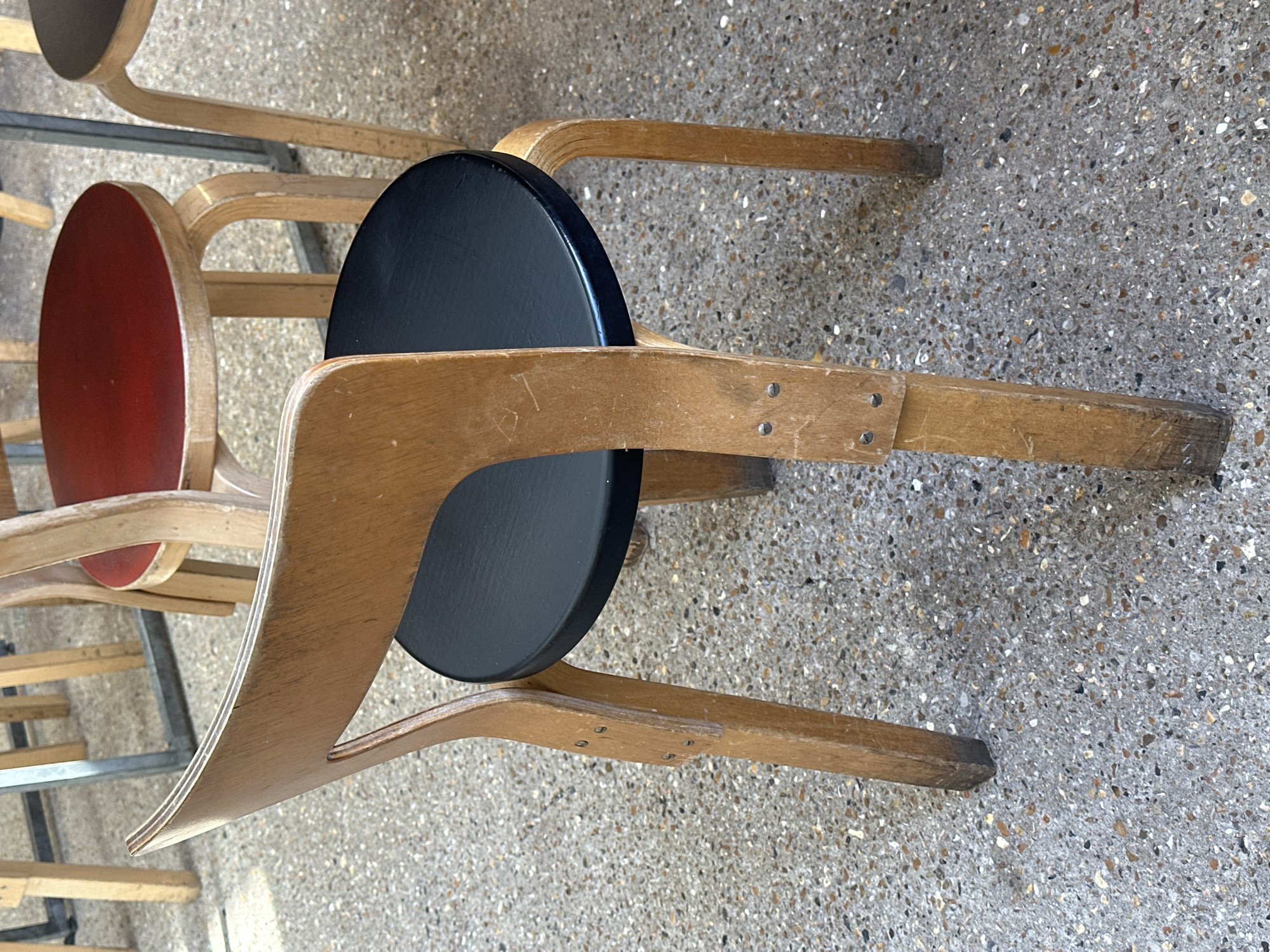 The image captures several wooden stools, likely stacked or closely arranged, set against a speckled concrete floor in London, United Kingdom. The stools feature a distinctive design with bent plywood frames and round seats. In the foreground, a stool with a black seat is prominent, while another behind it has a red seat. Other stools are visible further back, consistent with a utilitarian, possibly institutional or cafe, aesthetic.
The wooden components of the stools show clear signs of wear, including scratches, scuffs, and faded areas, suggesting frequent use. The bentwood construction is visible in the gracefully curved legs and supports, which are secured with metal screws. A faint inscription, possibly "BT" or "8T," is discernible scratched into the wooden frame of a stool near the black seat.
The setting appears to be an outdoor or semi-outdoor area, such as a patio or courtyard, indicated by the durable, exposed aggregate concrete flooring. The lighting is bright and even, characteristic of daytime. A metal rail or frame is partially visible in the background, possibly part of a cart or stacking system for the stools. No human activity is depicted; the scene focuses solely on the arrangement and condition of the furniture.