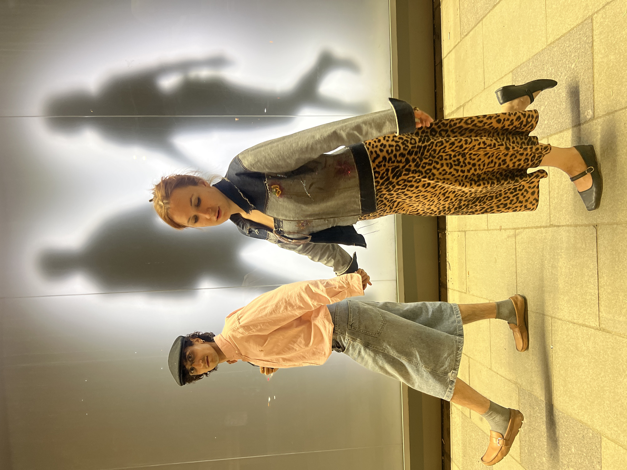 Two individuals stand in front of a reflective wall, casting shadows. The individual on the upper right wears a leopard print dress, a denim jacket, and black Mary Jane shoes. The individual on the lower left wears a pink button-up shirt, a denim skirt, a newsboy cap, brown shoes, and gray socks. They are standing on a tiled floor. It is plausible that this image was taken in Moscow, Russia, given the metadata, but no aspect of the visual image indicates that this is so.