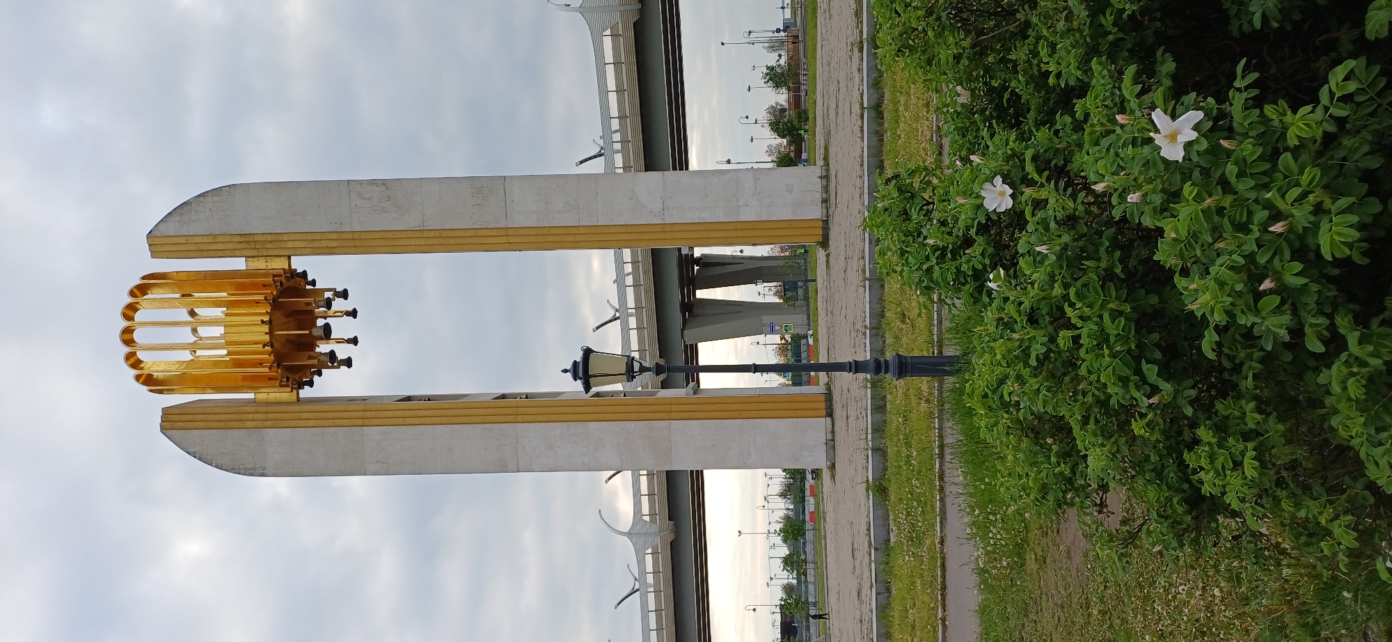 The image captures a scene near the foot of the Krestovsky Island Bridge in Saint Petersburg, Russia. A prominent architectural feature is a large, gray monument with gold trim and a decorative gold horn-like element attached to the upper portion. A vintage lamppost stands adjacent to the monument. In the background, the Krestovsky Island Bridge spans across the scene. Lush greenery with white flowers is present on the right side of the image. A paved pathway runs along the scene. The sky is overcast. The image is oriented sideways.