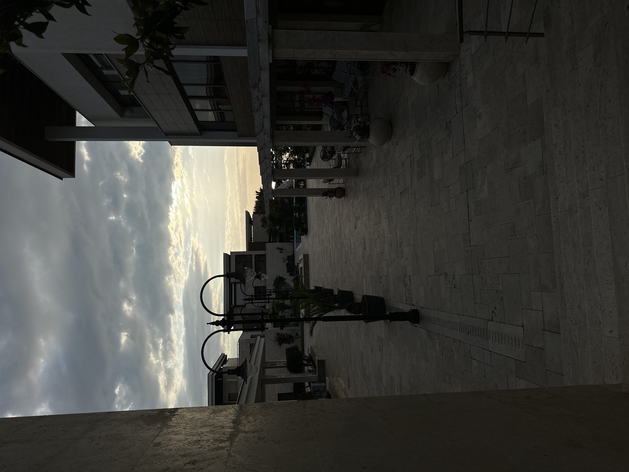 The image depicts an outdoor courtyard area in Sozopol, Bulgaria, likely a residential or hotel complex. The time of day appears to be late afternoon or early evening, with the sky overcast and the sun setting behind the clouds, casting a soft, diffused light. The courtyard is paved with large, square stone tiles.
In the foreground, a large stone pillar is visible on the left side, partially obscuring the view. To the right, a modern building with large windows and a dark facade is prominent. An outdoor seating area with white furniture and striped cushions is visible under a white pergola.
A row of decorative street lamps with arched tops lines the central pathway. Potted plants, including palm trees, are strategically placed along the pathway and near the buildings. In the background, more contemporary buildings with flat roofs are visible. A swimming pool can be seen in the distance, suggesting a leisure or resort setting. The overall atmosphere is tranquil and slightly subdued due to the lighting conditions. There are no people visible in the scene.
