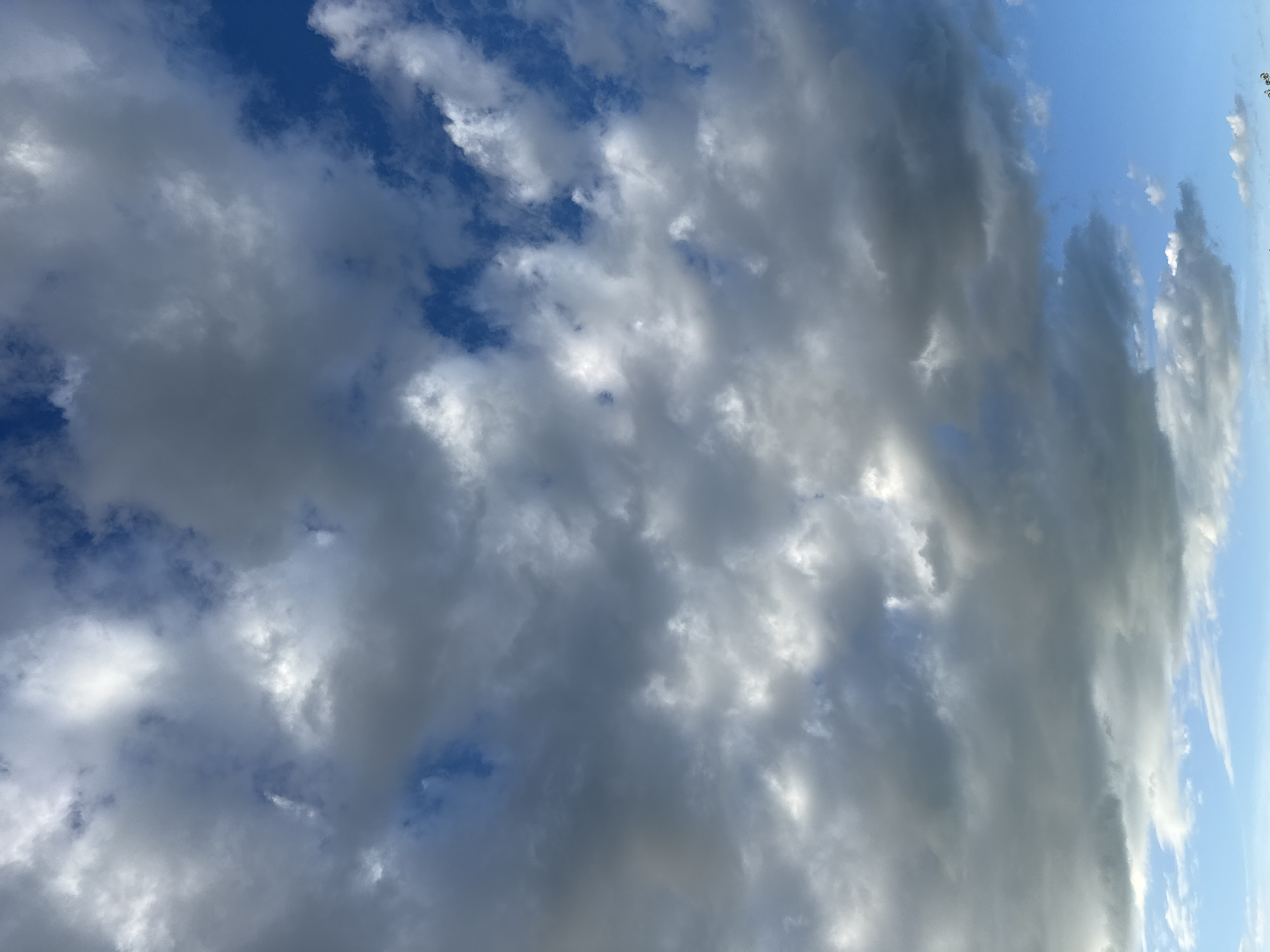 The image is a close-up, vertical shot of a cloudy sky. The clouds are a mix of white and dark grey, with patches of bright blue sky visible between them. The lighting suggests it could be late afternoon or early morning, with sunlight filtering through the clouds. There are no people, subjects, objects, actions, or interactions depicted in the image. The environment is solely the sky. The location in the city of Baan Chaweng, Thailand is not discernible from the visual information provided in the image.