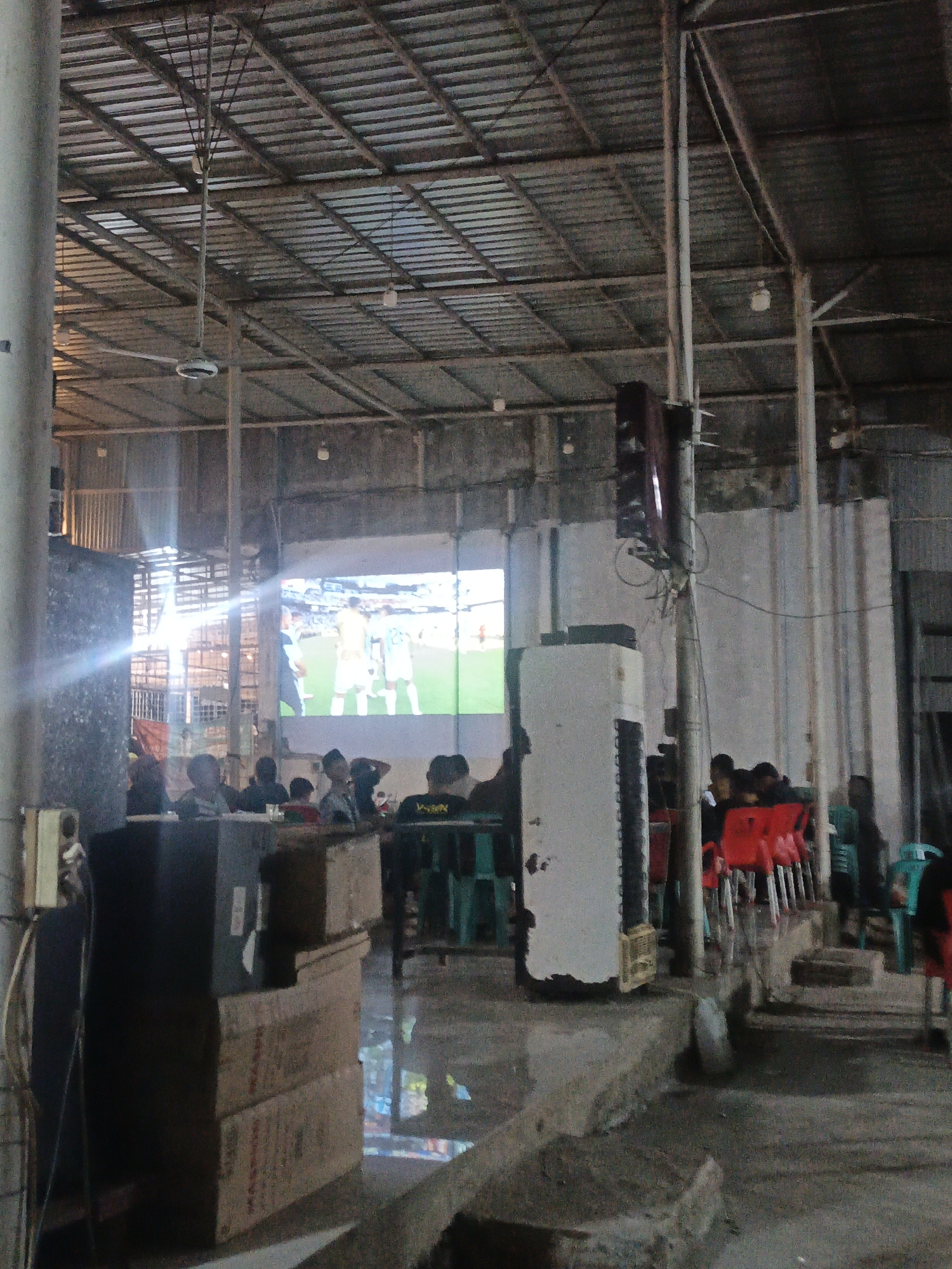 The image depicts an indoor gathering, possibly a sports bar or community hall, in Keudejungkagajah, Indonesia, where a group of people are watching a projection of a soccer match. The scene is dimly lit, with the bright projection screen serving as a primary light source. A large ceiling fan is visible, suggesting a warm environment. The setting appears to be rustic, with exposed metal roofing, concrete floors, and a mix of plastic and metal chairs. In the foreground, several stacked cardboard boxes are present, along with electrical conduits and a large, white, vertical appliance that may be an air conditioning unit or a projector housing. The attendees, mostly male, are seated and observing the projected image. The overall atmosphere is one of casual observation and communal viewing of a sporting event.