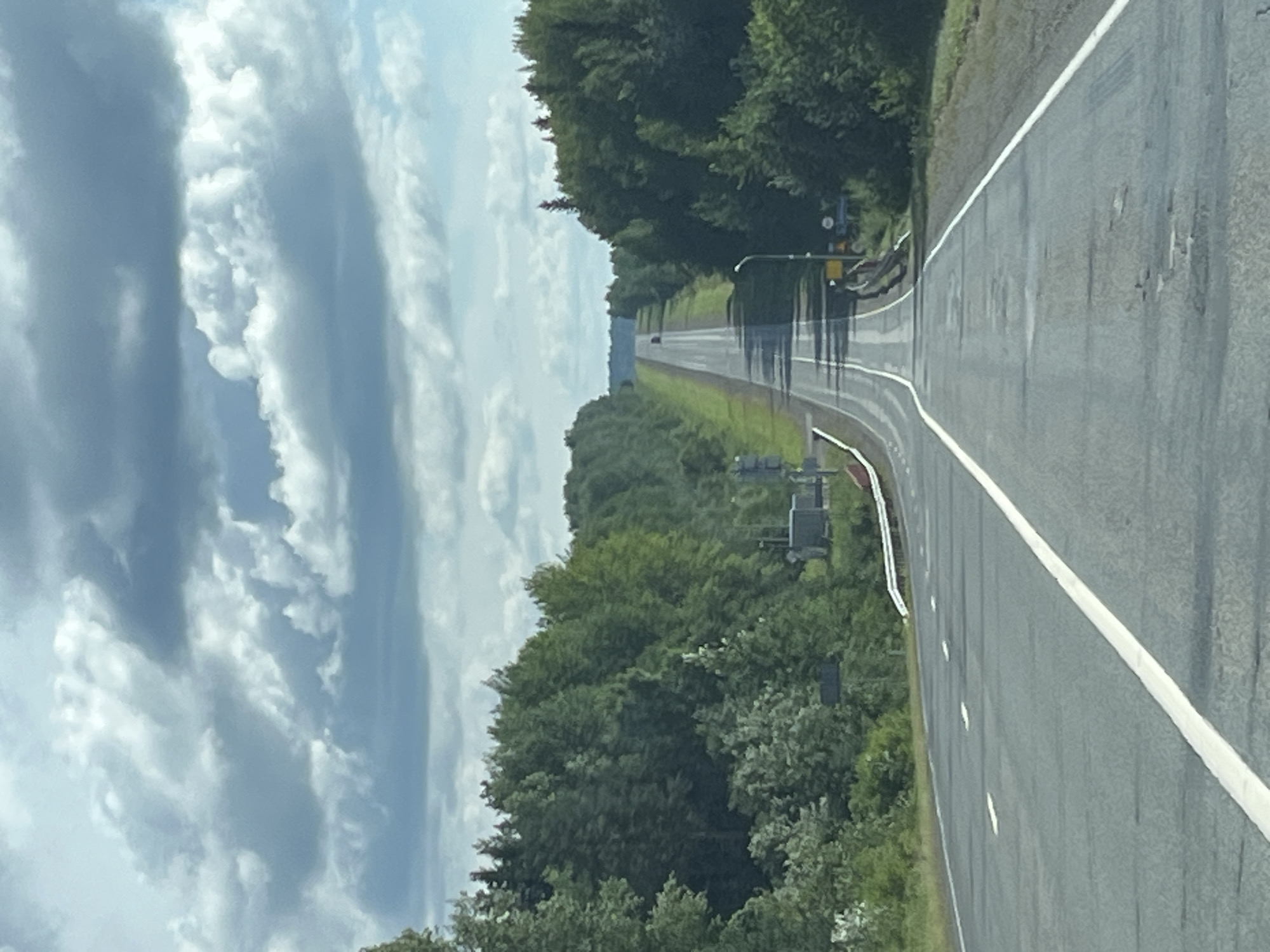 This is a photograph taken from inside a vehicle traveling on a two-lane asphalt road that curves gently and rises towards the horizon. The road is bordered by dense green trees on both sides, with a clear blue sky and scattered clouds above. In the distance, on the road, a dark-colored car is visible. The scene appears to be in a rural or suburban area, likely on the outskirts of Pal', Russia, given the prevalence of forested landscapes and the typical appearance of Russian roads. There are no people or specific landmarks clearly identifiable that would pinpoint the exact location within Pal'. The overall impression is one of a scenic drive through a wooded region under a partly cloudy sky.