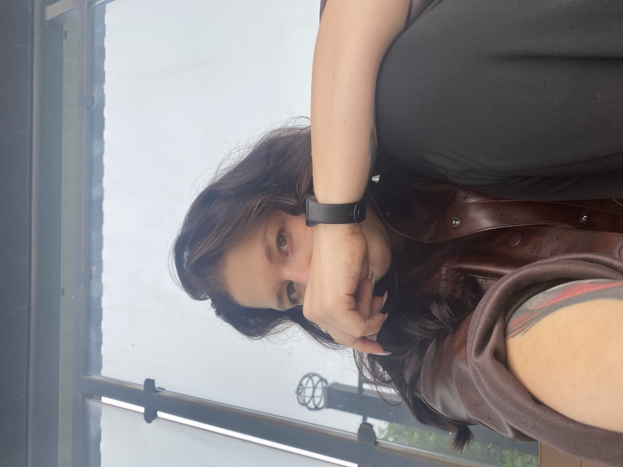 This vertically oriented close-up selfie features a person with long, dark, wavy hair and light-colored eyes, looking directly into the camera. Their left hand is raised, partially covering their mouth and chin. A black smart band or watch is worn on their left wrist, and a tattoo with red and black abstract lines is visible on their right forearm, partially obscured by a dark brown or maroon leather jacket. They are wearing a black top or dress underneath the jacket, and their nails are light-colored and appear manicured.
The person appears to be indoors, positioned next to a large window or glass partition. The background visible through the window is a bright, overcast sky, indicating a cloudy or diffused light day. Dark window frames are present on the left and bottom edges of the image. A reflection, or a view through the glass, shows a decorative metal sphere mounted on a post, suggesting an outdoor architectural feature. The overall lighting is soft and natural due to the overcast conditions. The person's expression is neutral, and the pose could be interpreted as casual or slightly pensive. No visible text is discernible in the image. The scene is set in Chaikovsky, Russia.