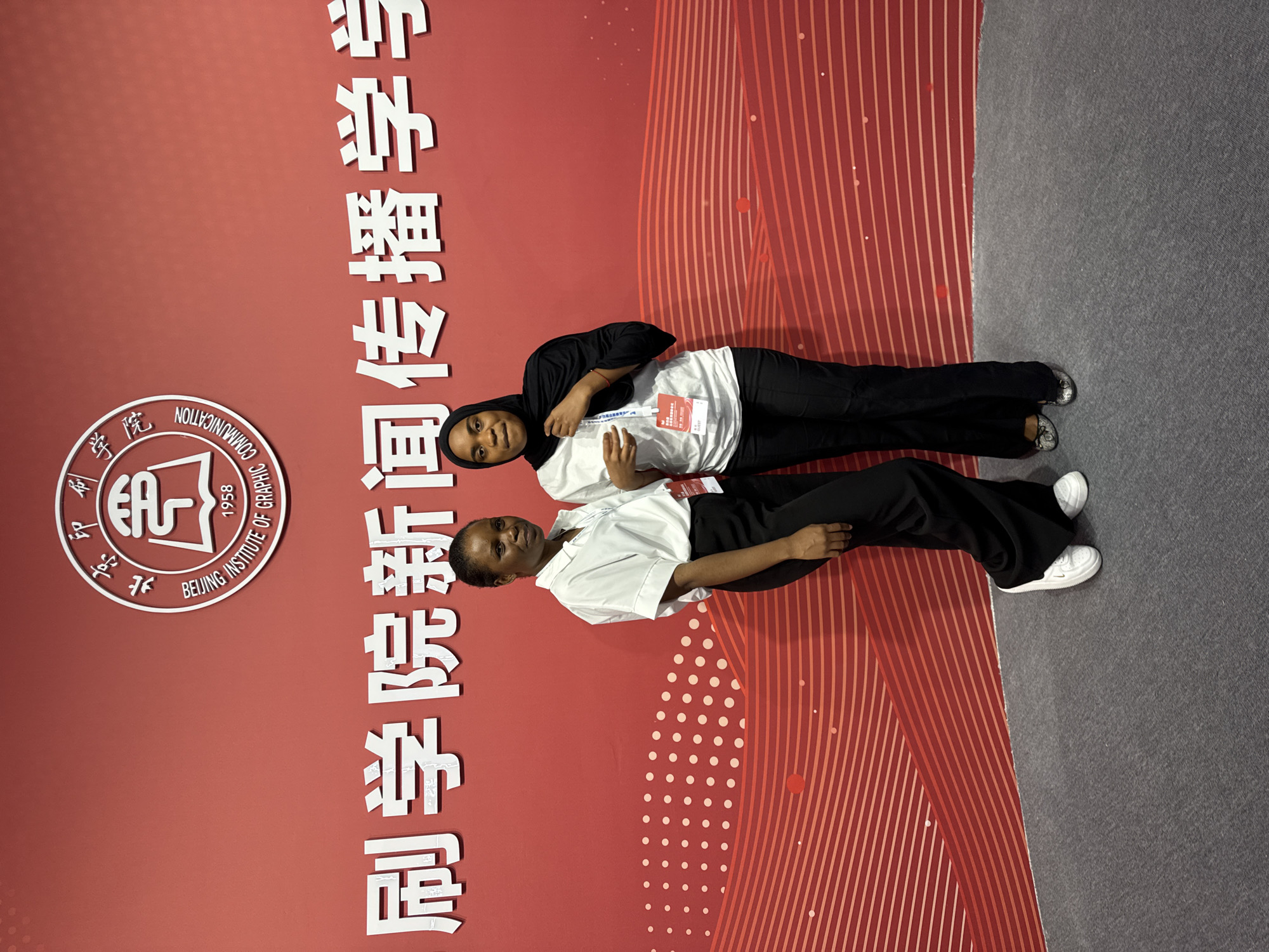 Two women stand in front of a red backdrop with Chinese characters and a circular logo. The woman on the left wears a white button-up shirt and black pants, her right foot is forward, and she looks towards the camera. The woman on the right wears a black hijab, a white top, and black pants. She holds her hijab with her left hand and looks slightly away from the camera. Both women have lanyards with identification cards around their necks. The backdrop features a repeating pattern of white dots and red wavy lines, with the text "BEIJING INSTITUTE OF GRAPHIC COMMUNICATION" encircling the logo. The floor is carpeted in a dark gray color. The overall scene suggests an event or presentation at the institute. The location is the Daxing District, China.