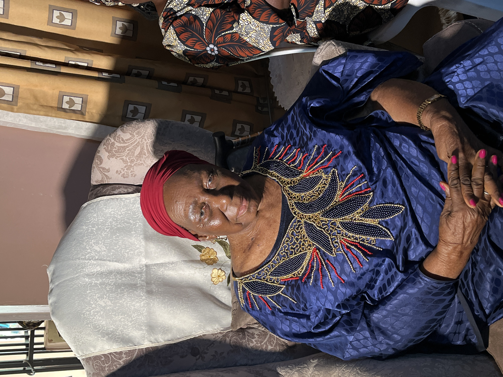 A medium shot shows an elderly African woman with dark skin and a red headscarf seated in a chair. She is wearing a dark blue, patterned dress with intricate gold and red beadwork around the neckline. Her hands are clasped in her lap, and she is wearing a gold bracelet on her left wrist. Her fingernails are painted bright pink. Her face is turned slightly towards the camera, and she is smiling. In the background, to the right, a portion of another person in a patterned brown and orange dress is visible. Behind the woman, there are textured curtains and a light-colored decorative cloth draped over the back of the chair. The scene appears to be an indoor setting, likely a home. The image was taken in Bukuru, Nigeria.