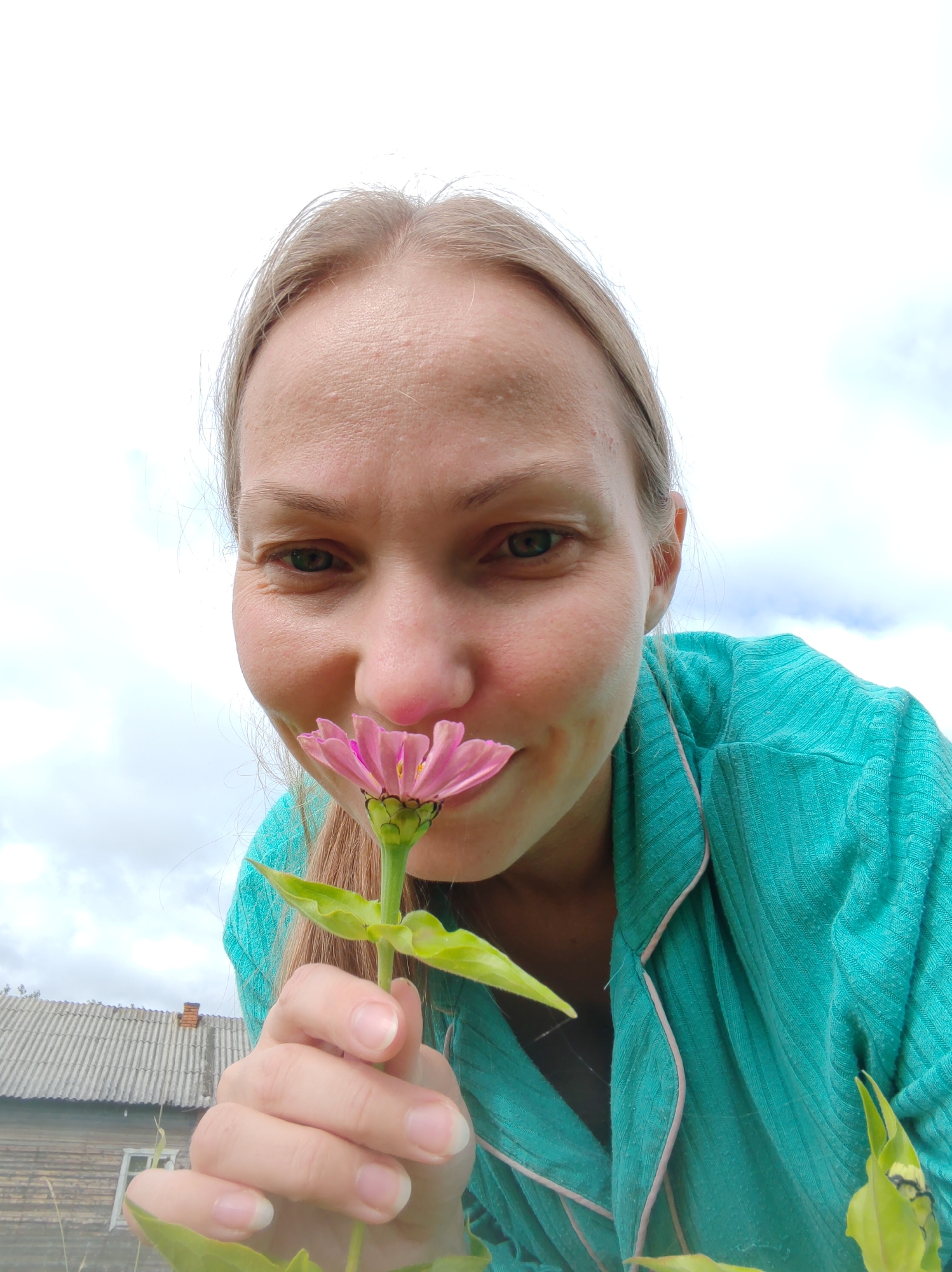 A young woman with blonde hair is shown from a low angle, holding a pink flower to her nose and smelling it. She is wearing a teal-colored top with pink trim. In the background, there is a wooden building with a corrugated roof and a small window. The sky is overcast with clouds. The image appears to have been taken outdoors in a rural setting, likely in or near the city of Chukhlom, Russia.