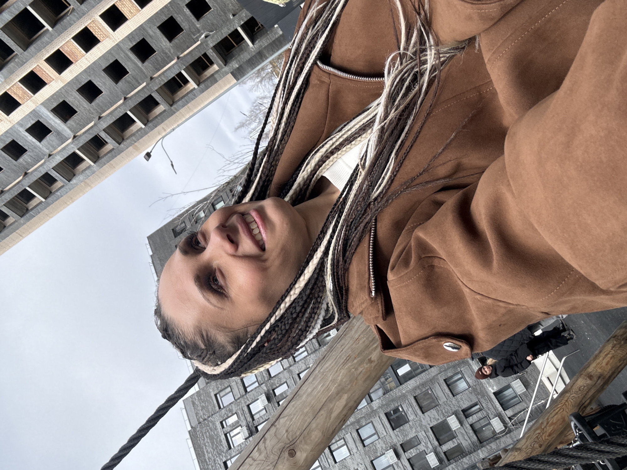 The image is a low-angle selfie featuring a woman with brown and white braided hair, wearing a brown jacket over a white top. She is smiling slightly. The background consists of multi-story, grey brick buildings with numerous window openings, suggesting a modern urban environment. In the lower part of the image, there are sections of wooden and rope structures. Two people, dressed in dark clothing, are visible at the lower right side of the image. The scene implies an outdoor location, likely a pedestrian walkway or bridge, set against the backdrop of apartment buildings in the Rusanovo district of Russia.