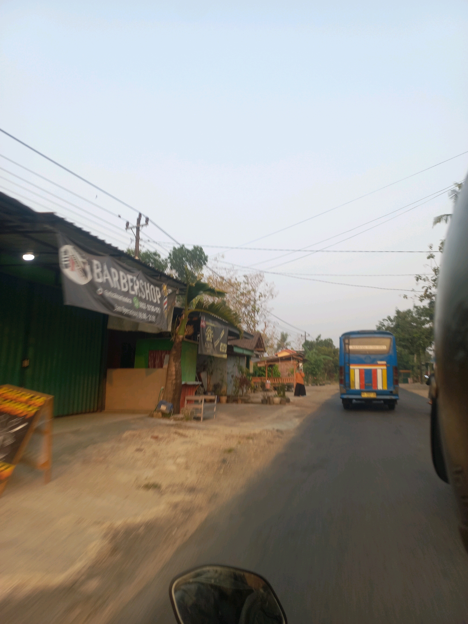 The image depicts a street scene in Ngarjosari, Indonesia, likely during the late afternoon or early evening due to the soft, diffused light. In the foreground, a dirt or unpaved road occupies the lower portion of the frame, with a blurred reflection of a helmet mirror on the right side. A blue bus with yellow, red, and white stripes on its rear is traveling away from the camera on the paved road. To the left of the road, a barbershop is visible with a dark banner advertising "BARBERSHOP" and showing a phone number, suggesting a commercial establishment. Green metal doors are part of the barbershop's facade. Adjacent to the barbershop, there are small buildings and some trees. A person in an orange garment is standing near the side of the road in the distance. Overhead, power lines crisscross the pale, hazy sky. The overall impression is of a modest, somewhat rural or suburban area.