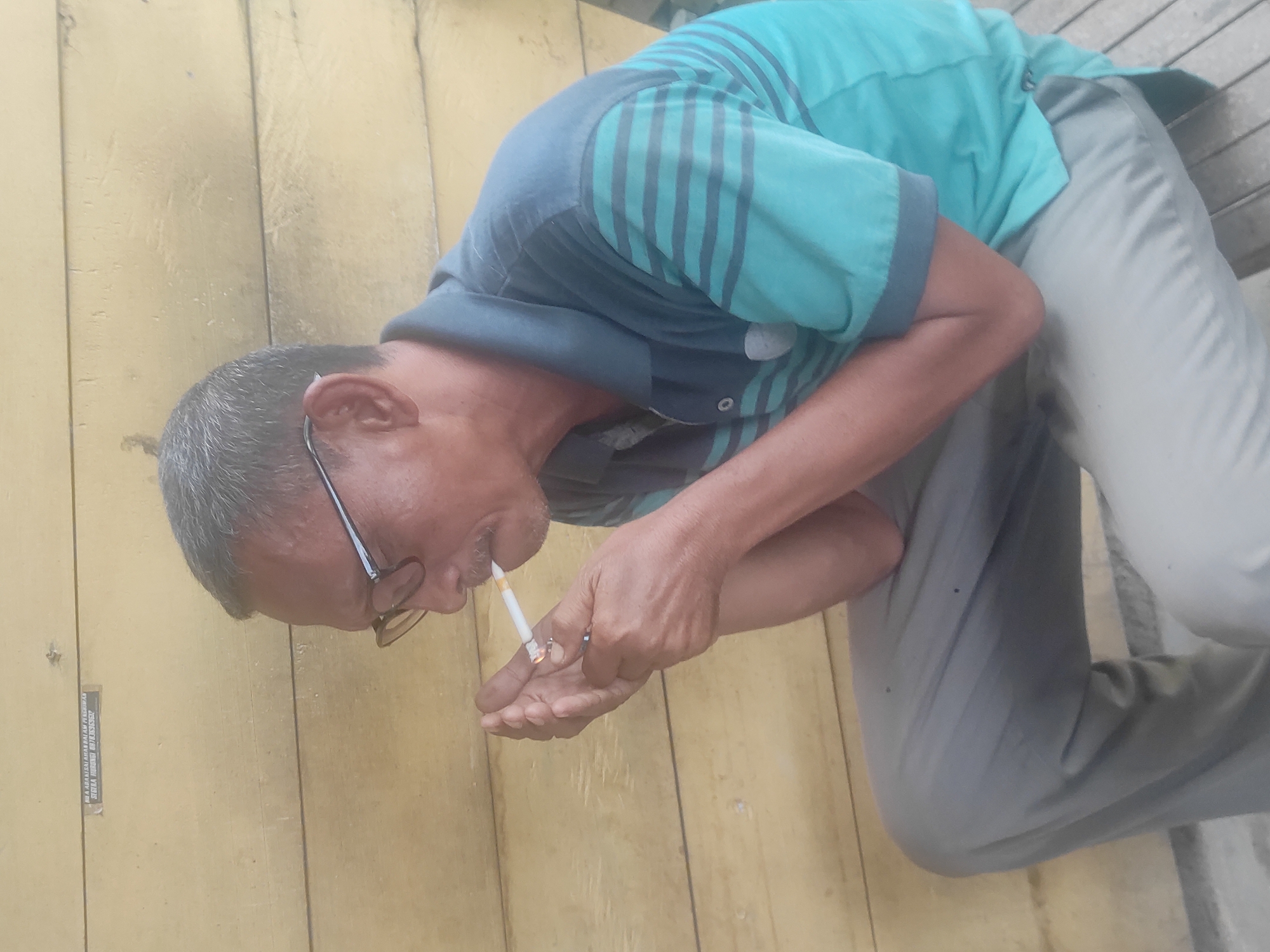 An Asian man in glasses, wearing a striped aqua-green and navy polo shirt and gray trousers, is bent over, lighting a cigarette with a lighter held in his left hand. He is outside on what appears to be a wooden platform or floor with an adjacent wall made of wood planks. A small label with the words "JIKA ANDA MERASAKAN PERTUMBUHAN SEGLA HUBUNGI 081361368502" is on the wooden wall. The location is in the city of Lhokseumawe, Indonesia.