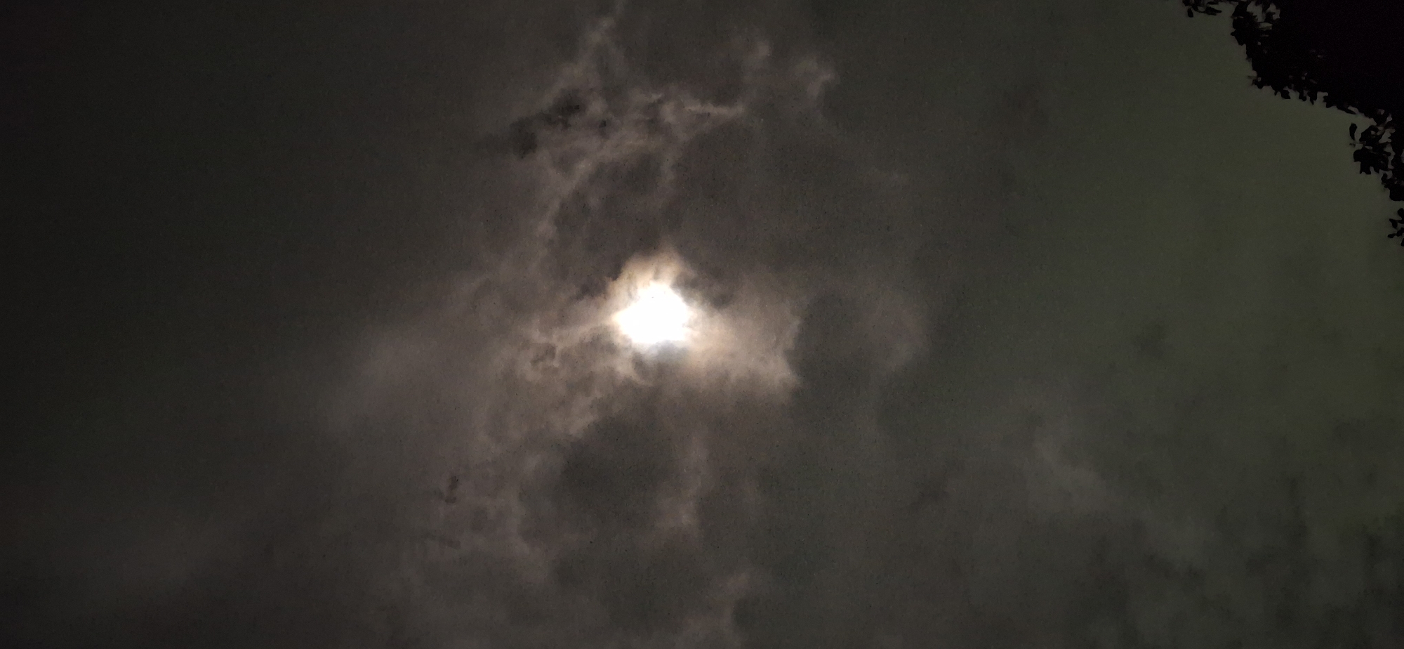 The image depicts a night sky in Vallecito, Venezuela. Heavy, dark clouds dominate the scene. A bright light source, obscured by a cloud, is visible in the center of the image. The dark outlines of tree branches are visible in the upper right corner of the image.
