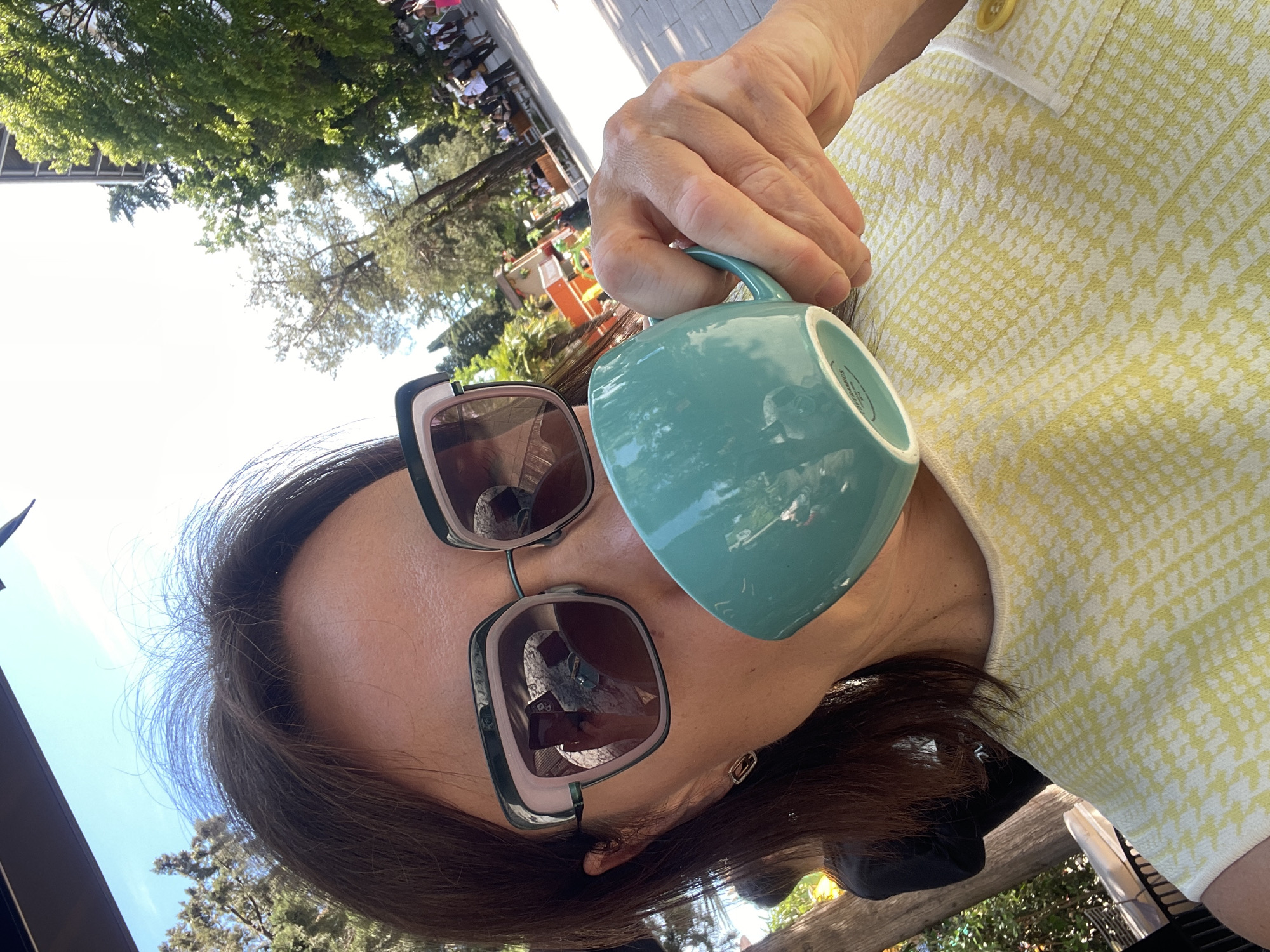The image, rotated 90 degrees clockwise, captures a close-up of a person outdoors on a sunny day in Sochi, Russia. The individual, likely an adult female, is partially visible, seen from her chin up, with dark hair framing her face. She wears large, stylish sunglasses featuring dark brown lenses and thick frames with light-colored accents.
She is dressed in a yellow and white patterned top, possibly a houndstooth or similar small geometric design, with a distinct white trim and a yellow button visible near her right shoulder. Her right hand holds a teal-colored ceramic mug, positioned near her mouth as if she is about to drink or has just taken a sip. The bottom of the mug features small, illegible text, which appears to include care instructions like "DISHWASHER SAFE" and "MICROWAVE SAFE."
The background reveals a vibrant outdoor setting under a clear, bright sky. Lush green trees, indicative of a park or garden, are prominent. In the mid-ground, blurred figures of people are seen seated at tables, suggesting a cafe or outdoor dining area. The bright lighting and visible foliage point to a warm, clear day, likely during the morning or afternoon. The scene conveys a sense of leisure and enjoyment of the outdoor environment.