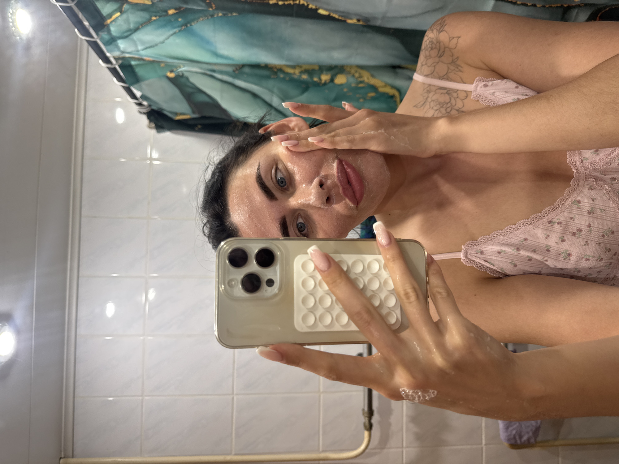 A woman is taking a selfie in what appears to be a bathroom. She has a facial cleanser on her face and is wearing a pink floral top. She is holding a smartphone in her left hand. The bathroom has white tile walls, a shower curtain with a blue and green abstract pattern, and some plumbing fixtures. The specific location is not discernible.