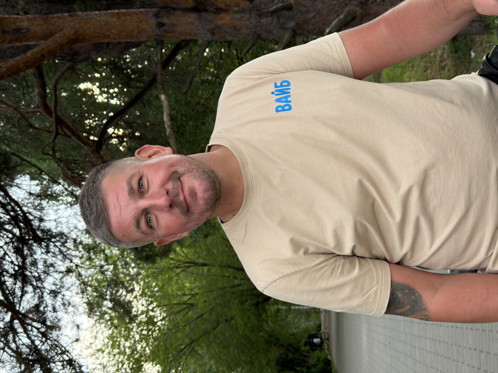 This is a close-up, portrait-style photograph of a man outdoors, with the camera angled to be nearly sideways, as if the photographer is lying down or the camera is tilted. The man is in the foreground, looking towards the camera with a slight smile. He has short, graying hair and a beard, and his eyes are blue. He is wearing a tan, long-sleeved t-shirt. On the upper left side of his shirt, there is blue text that appears to read "ВАЙБ" in Cyrillic. His left arm is visible, showing a tattoo on his forearm.
The background is out of focus, with dappled sunlight filtering through green foliage and tree branches, suggesting a wooded or park-like setting. The lighting indicates it is daytime, likely late afternoon or early evening, given the soft, warm quality of the light. The overall mood of the image is casual and relaxed. The location context of Chaikovsky, Russia, places the scene within a natural environment, possibly a forest or park common in that region. The man's attire and the natural backdrop suggest a leisurely outdoor activity.