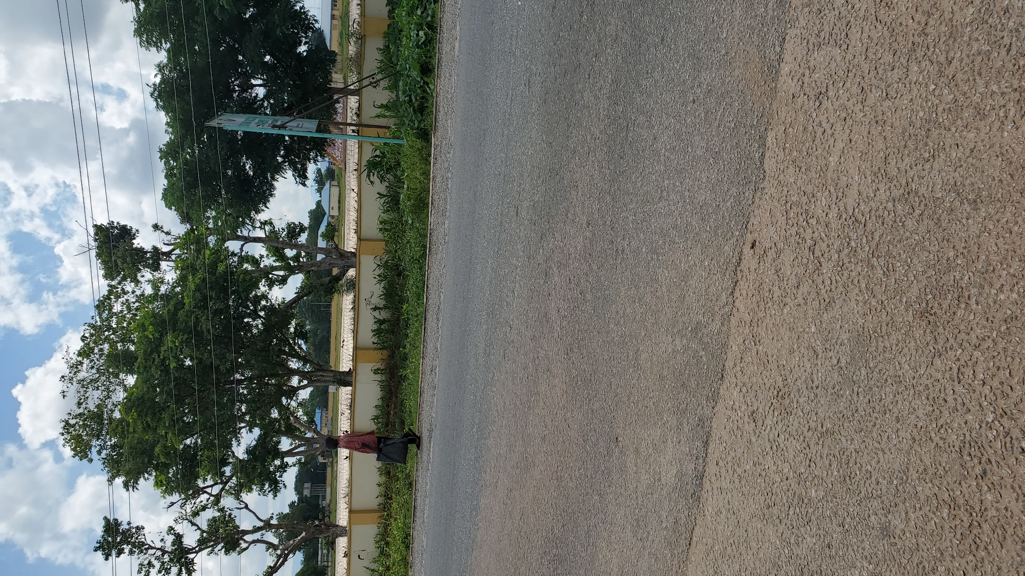 A lone person walks on the side of a paved road in Jalingo, Nigeria, carrying a dark bag. The person is wearing a patterned shirt and dark pants. The road has two distinct sections: a darker asphalt area in the foreground and a lighter, gravel-like area further back. Behind the road, a low yellow wall with barbed wire runs horizontally. Beyond the wall, a row of green bushes and scattered trees are visible, with some buildings and a blue water tower in the distance. The sky is partly cloudy with white cumulus clouds against a blue backdrop. A faint sign is visible on a pole in the background. The scene appears to be in an urban or semi-urban setting.