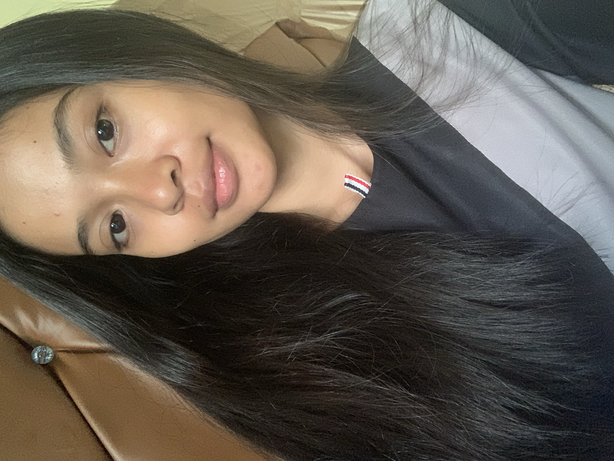 A young woman with dark hair is shown from the chest up, looking into the camera. She is wearing a black t-shirt with a grey diagonal stripe. She has natural makeup on, with a touch of lip gloss. Behind her, to the left, is a brown headboard with a decorative button. To the right, the background is out of focus, appearing to be a neutral colored curtain. The lighting is soft and natural, coming from the front. There is no visible indication of the woman's location in Sungai Meriam, Indonesia.