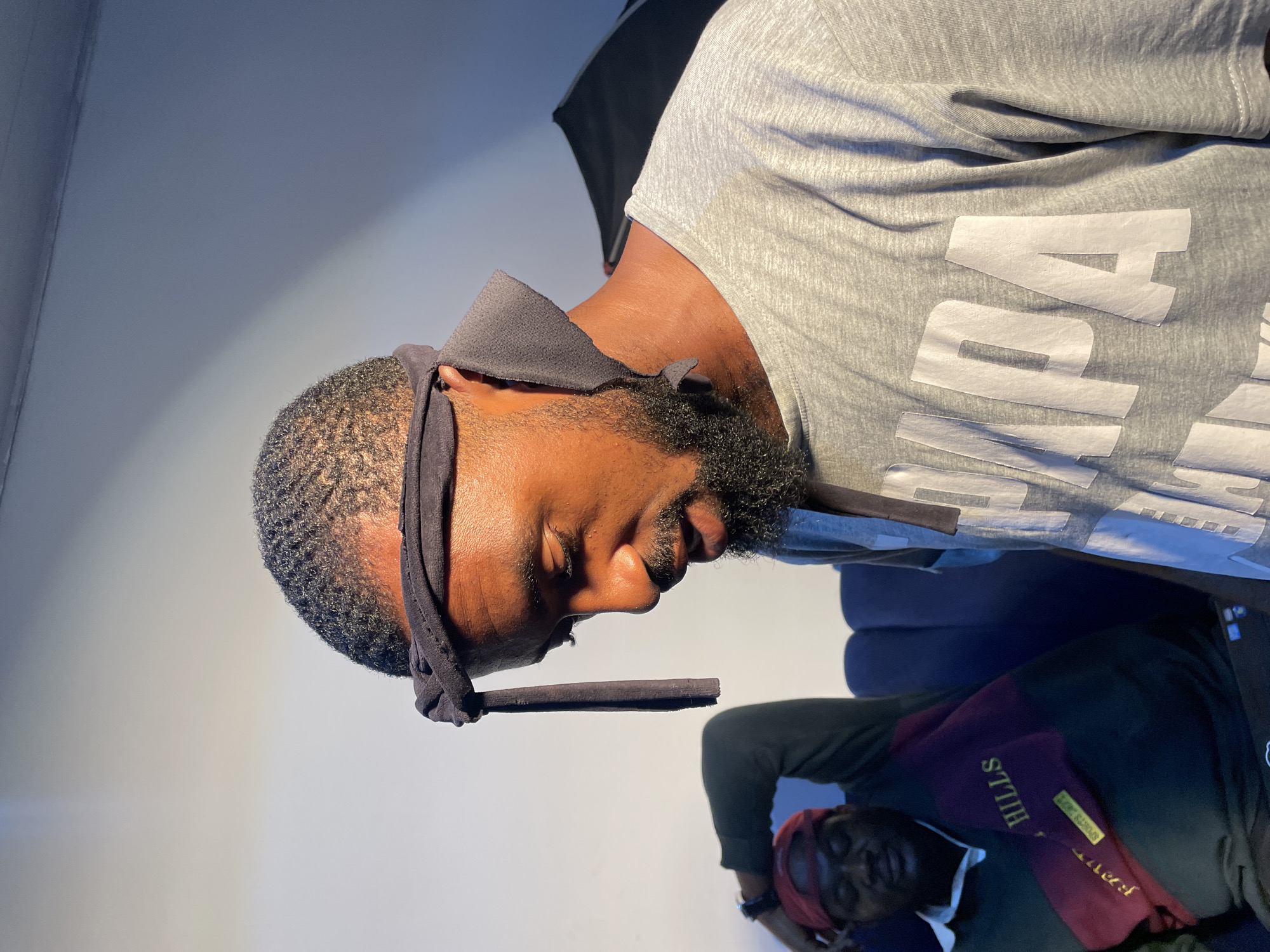 Two Black men are present in the image. One is wearing a grey t-shirt with white lettering. He has a black headscarf and a dark beard. A dark cylindrical object is affixed to the front of the headscarf, possibly a cigar or blunt. The second man is wearing a green shirt with "HILLS" partially visible on the front and a red headscarf. The location is possibly a photography studio, with a white wall in the background. There are no other contextual clues to place the subjects in Barakin Akawo, Nigeria.