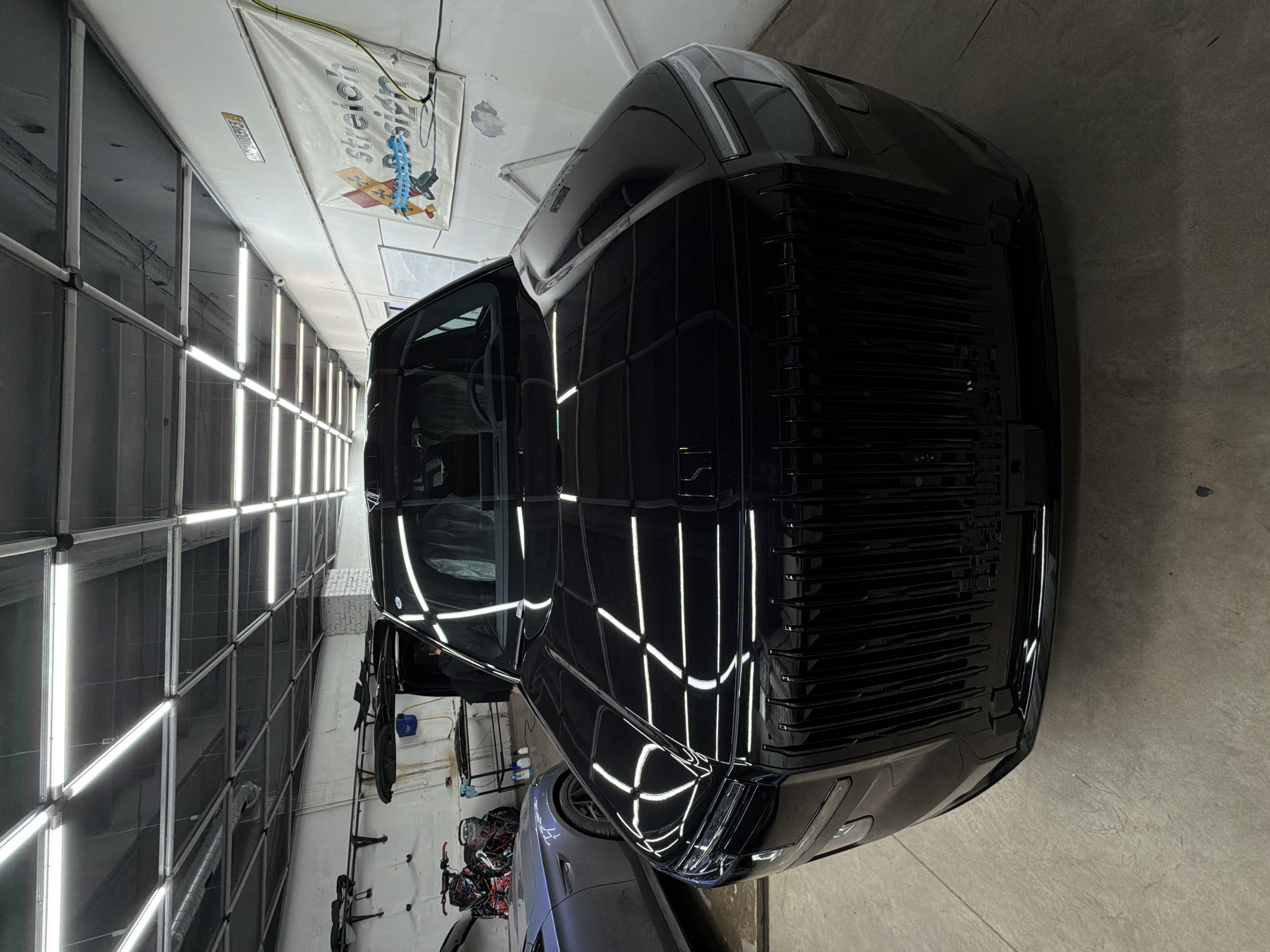 The image shows the front of a black vehicle, possibly a large SUV or van, parked inside a garage or workshop. The location seems to be an automotive detailing or repair shop, as indicated by the overhead lighting grid, the "stretch design" banner on the wall, and the other vehicles and equipment in the background. Additional objects in the background include bicycles, tools, and cleaning supplies. The image does not show evidence confirming the location in the city of Krasnoyarsk, Russia.