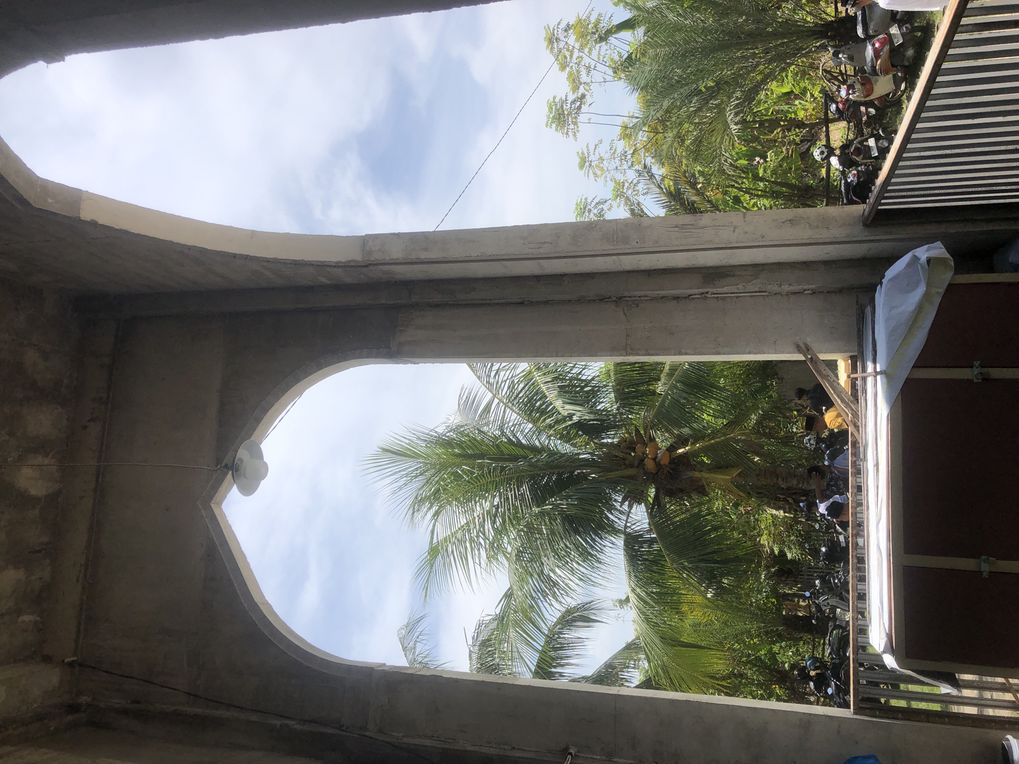 This image captures a view through an arched entryway in Lhokseumawe, Indonesia. The scene is bathed in daylight under a partially cloudy sky. In the foreground, concrete pillars and arches frame the view. A light fixture hangs from the ceiling of the entryway. Beyond the structure, lush green palm trees and other tropical vegetation dominate the landscape. Several motorcycles are parked in the middle ground, suggesting a gathering or a public area. A few individuals are visible, some near the motorcycles and others further in the background, their activities not clearly discernible. A wooden structure covered with white fabric is positioned in the lower part of the frame, partially obscured by a metal railing. The overall impression is that of a tropical, possibly public, outdoor space.