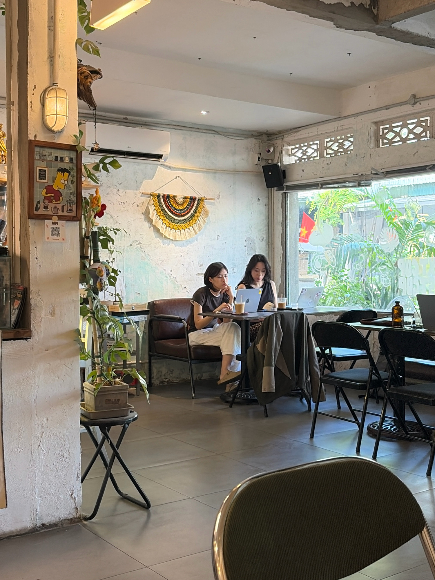 The image depicts an indoor scene at a cafe in Da Nang, Vietnam. Two young women are seated at a table by a large window, both engaged with laptops. The woman on the left is wearing a dark t-shirt and white pants, while the woman on the right is wearing a dark, sleeveless top and pants. In front of them are drinks and their laptops. The cafe's interior features a rustic aesthetic with exposed concrete walls, a tiled floor, and a decorative wall hanging. Plants are visible, including a tall potted plant on a small black folding stool near the left side of the frame. A vintage-style light fixture and framed art are also present on the wall. Outside the window, lush greenery and a Vietnamese flag are visible. Several chairs are arranged around other tables in the cafe.