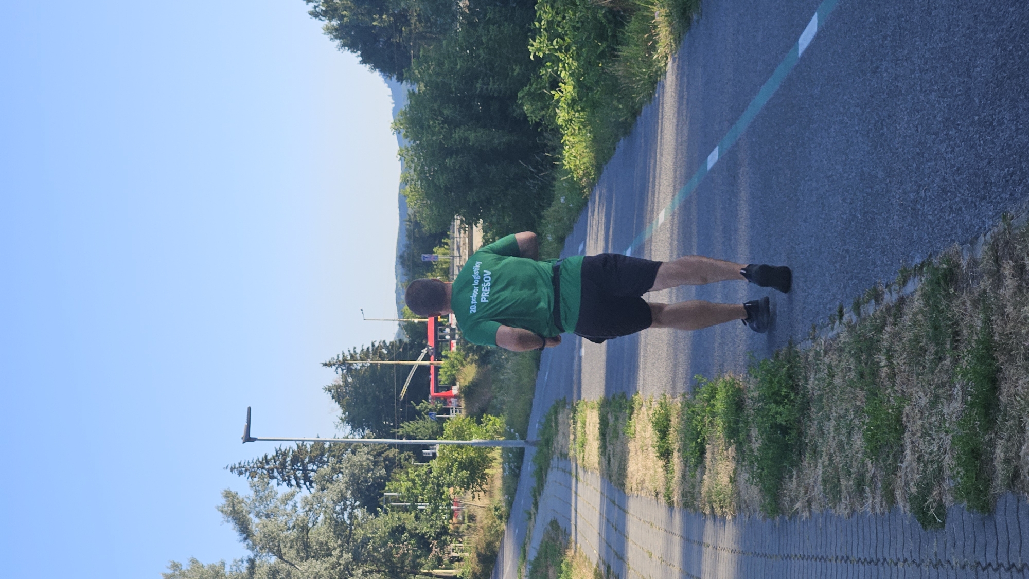 A man is jogging on a paved path next to a sidewalk in Prešov, Slovakia. He is wearing a green t-shirt with white text on the back that reads "20.príprav logistiky PREŠOV" and black shorts. The path has a white and blue line marking its edge. The environment includes trees, grass, a street lamp, and a red trolley bus in the background. The sky is clear and blue.