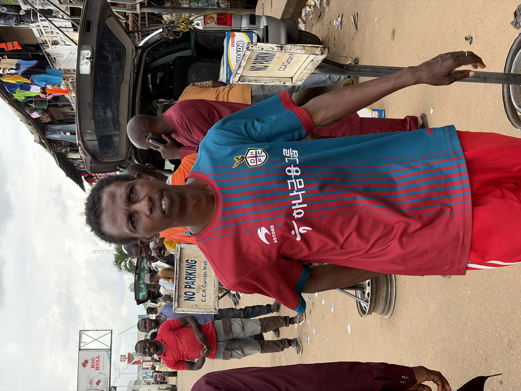 The image depicts an outdoor scene in Jos, Nigeria, likely a market or roadside area. In the foreground, a man with dark skin and short, dark hair is looking towards the camera. He is wearing a short-sleeved shirt with vertical stripes in red and teal, featuring a crest and Korean text on the chest. He also wears red shorts with white stripes on the sides. His right arm is visible, and he is wearing a ring on his right hand.
Behind him, another man in a red polo shirt and grey pants is partially visible. Several other people are present in the background, some appearing to be engaged in conversation or looking around. A silver SUV with its rear hatch open is parked in the middle ground. Clothing items are displayed on a rack above a stall to the right. Two "NO PARKING" signs are visible, one in the foreground near the main subject and another further back. The ground is paved with dirt and scattered debris. The sky is overcast.