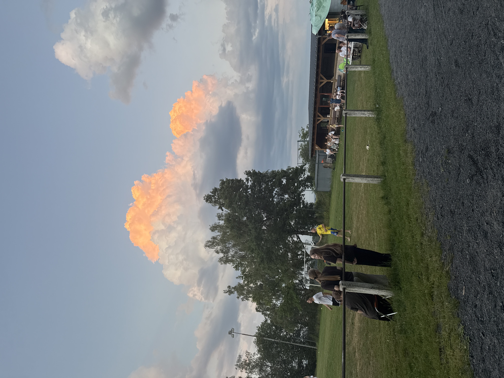 This is a forensic, matter-of-fact description of an outdoor scene in Atzenhain, Germany, at dusk. The foreground is a gravel path. Behind it, a grassy field with a fence runs horizontally across the image. Beyond the fence, several people are scattered across the field. In the midground, a group of trees stands tall, and a soccer goal is visible. A building with a covered outdoor seating area and tables is situated in the background. The sky above is filled with dramatic clouds, some of which are illuminated with an orange glow, suggesting the setting sun. The overall atmosphere is relaxed, with people engaged in various activities like playing soccer and socializing.