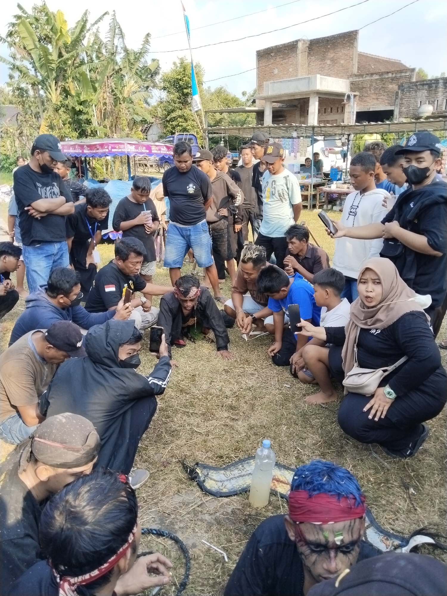 This image captures an outdoor gathering of people, primarily men, in Kedawung, Indonesia, during what appears to be a local event or performance. The scene is set on a dry, grassy field under bright daylight with a clear sky. In the background, lush banana trees and other foliage are visible, along with simple brick buildings and what might be vendor stalls covered with blue tarpaulins. A blue and white flag flies from a pole in the distance.
A large crowd, mostly young to middle-aged men, is congregated, forming a rough circle around a central activity happening on the ground. Many individuals are standing, while a significant number are crouching or sitting on the grass, observing with keen interest. Several people are holding up mobile phones, actively recording or photographing the event.
At the heart of the gathering, several men are on the ground. One man, wearing a black long-sleeved shirt, is on his hands and knees, seemingly a central figure in the activity. Surrounding him, others crouch, some also holding phones. In the foreground, two men stand out: one has vibrant blue hair, a red headband, and elaborate black and green face paint, displaying a focused expression. Another man next to him wears a traditional dark head covering. Many of the participants on the ground are barefoot.
On the right side of the frame, a woman wearing a brown hijab and a black top squats, looking intently towards the center of the activity, her mouth slightly open, suggesting surprise or engagement. She carries a light-colored shoulder bag. Near the blue-haired man, a plastic bottle containing yellowish liquid and a coiled dark rope are visible on the grass.
In the mid-ground, behind the crowd, parts of blue-tarped structures are visible. On one of these, faint text can be discerned, repeatedly mentioning "BENAMAKAN AZKEYLA PUTRI," suggesting names or perhaps a small business operating from the stalls. The overall atmosphere conveys a community-focused event, likely a cultural performance or a demonstration of some kind, drawing concentrated attention from the attendees.